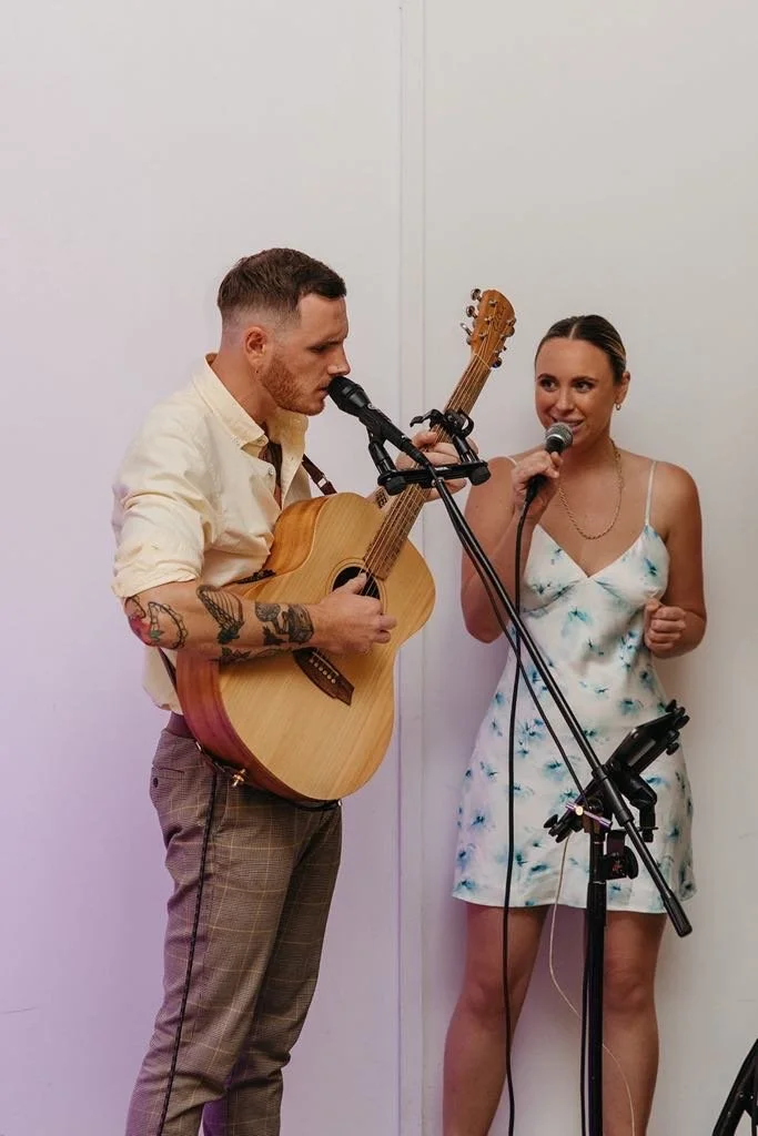 A man playing an acoustic guitar and singing into a microphone while a woman stands next to him, holding a microphone and singing, in a plain white room.