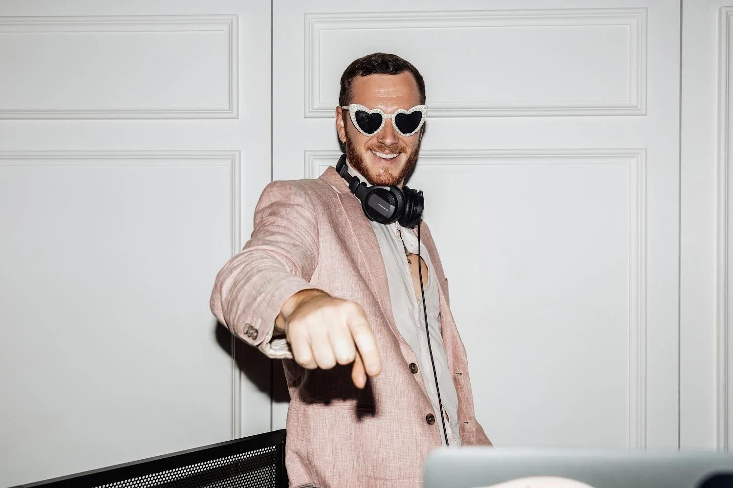A man wearing heart-shaped sunglasses, a pink blazer, and headphones around his neck, smiling and pointing towards the camera against a white paneled wall background.