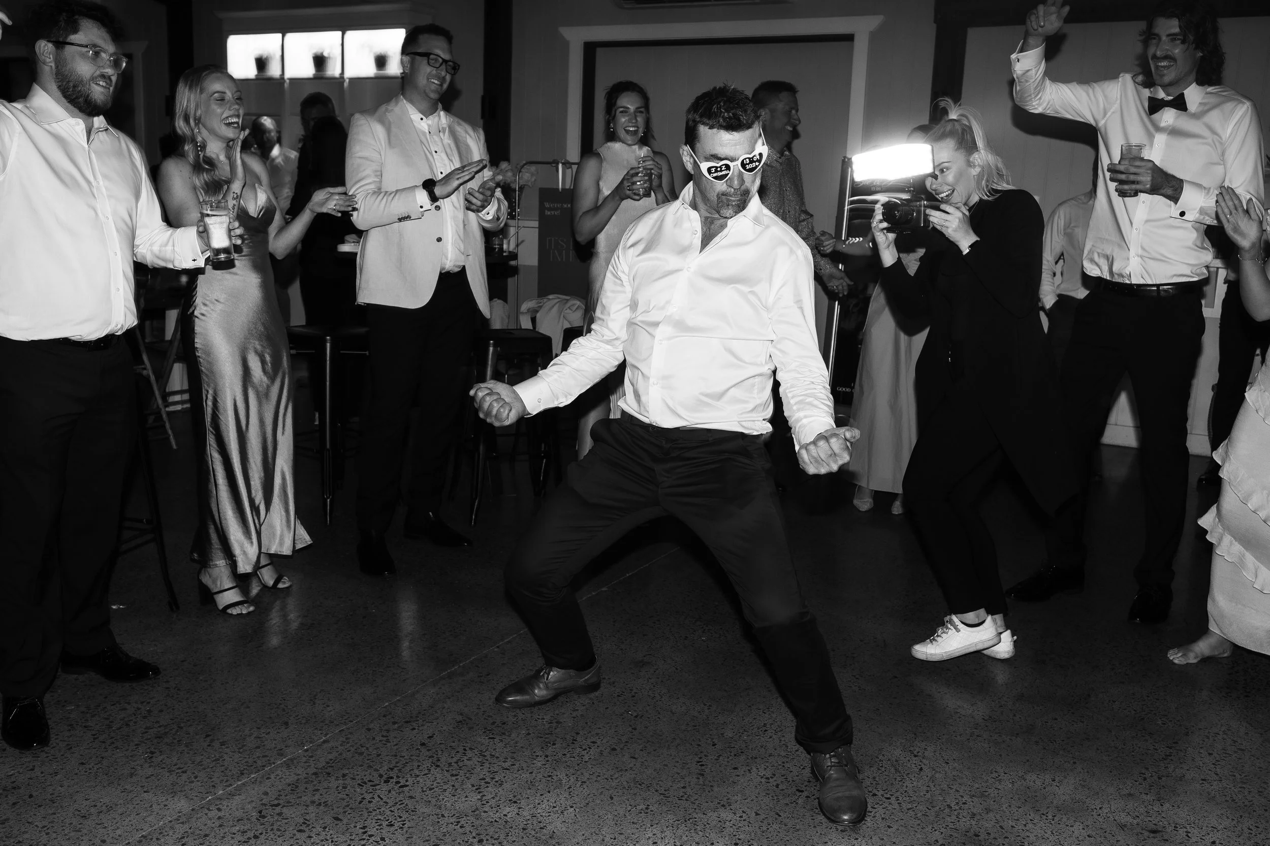 Group of people at a celebration or party, some holding drinks, others laughing and cheering. One man in the center, wearing sunglasses and a white shirt, is dancing or posing energetically. The scene is lively and joyful.