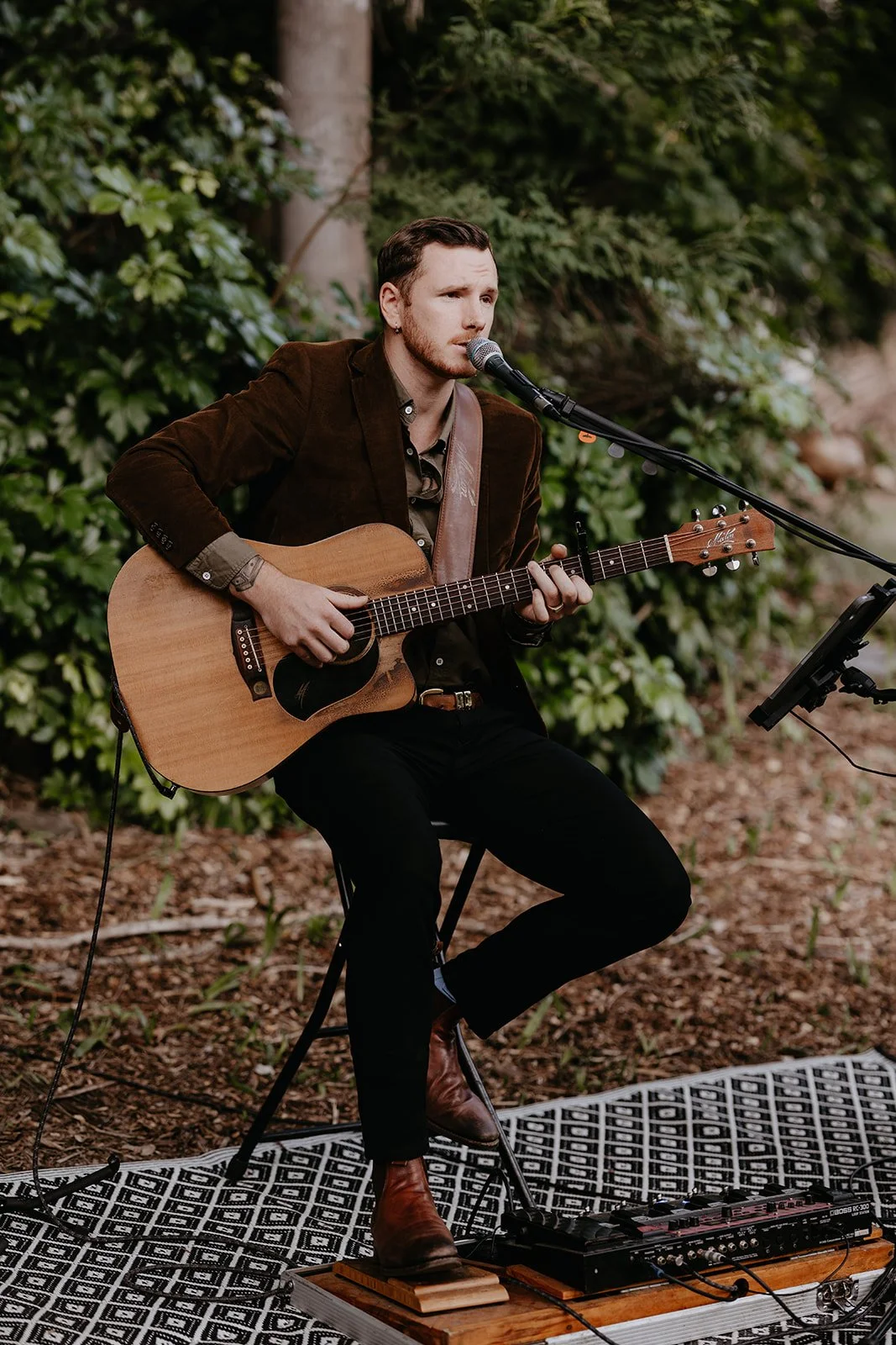 A man playing an acoustic guitar and singing into a microphone outdoors surrounded by greenery.