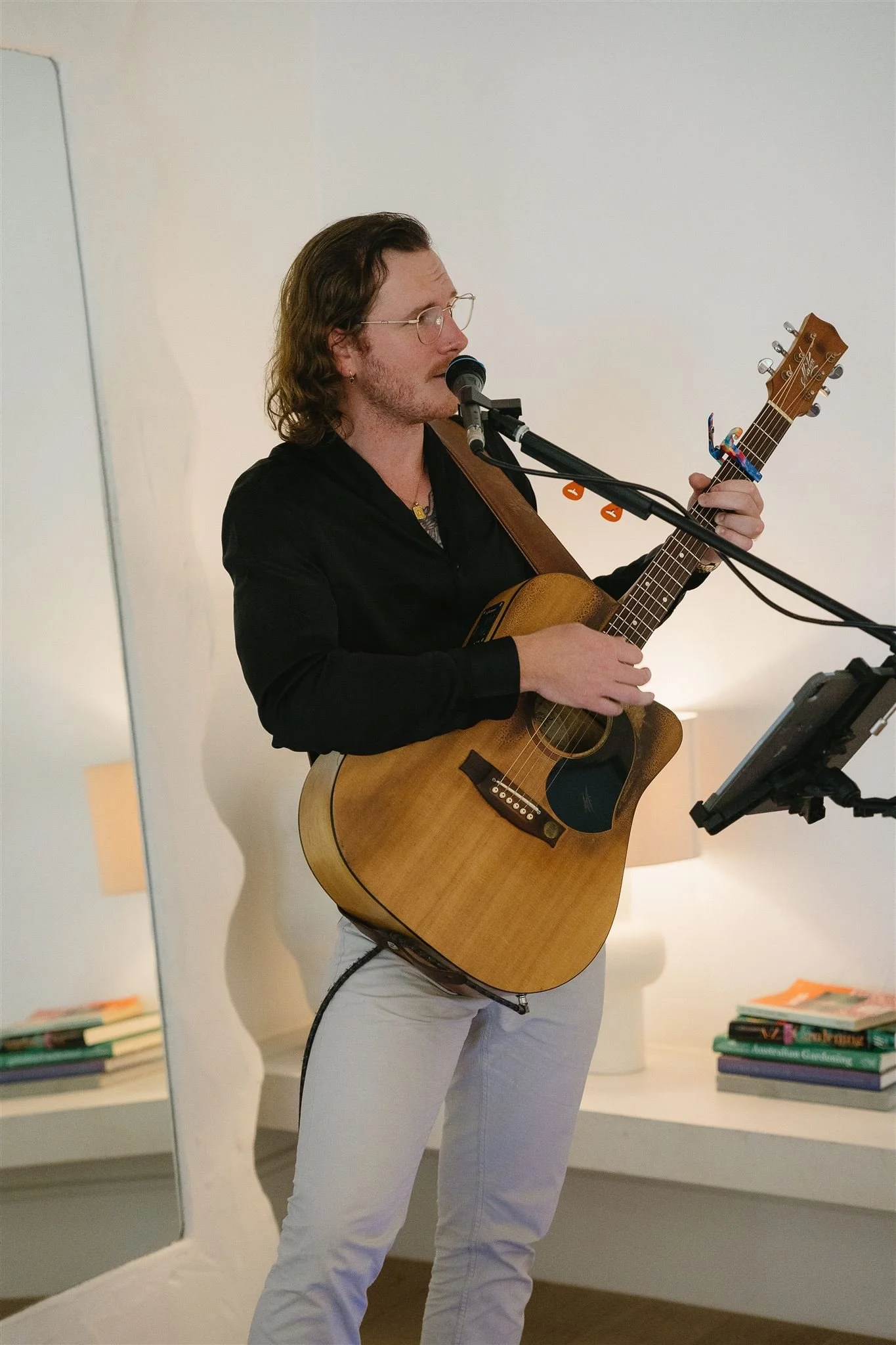 A person with long, wavy brown hair, glasses, and a small earring is playing an acoustic guitar and singing into a microphone. They are wearing a black shirt and white pants, and are standing indoors near a mirror and a bookshelf.