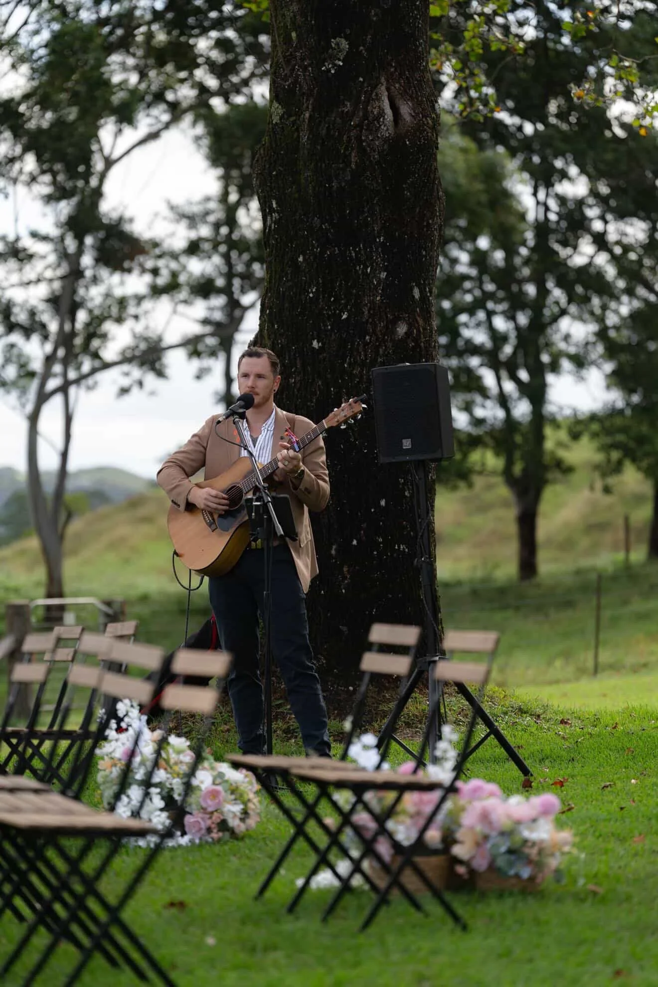 A man in a beige blazer playing guitar and singing into a microphone outdoors, with a large tree behind him and chairs with flowers in front.