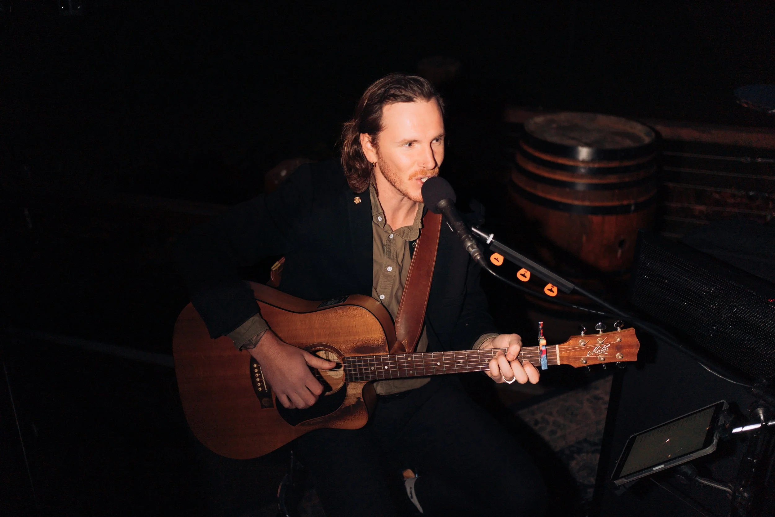 A man with light skin and long brown hair playing an acoustic guitar and singing into a microphone in a dark room