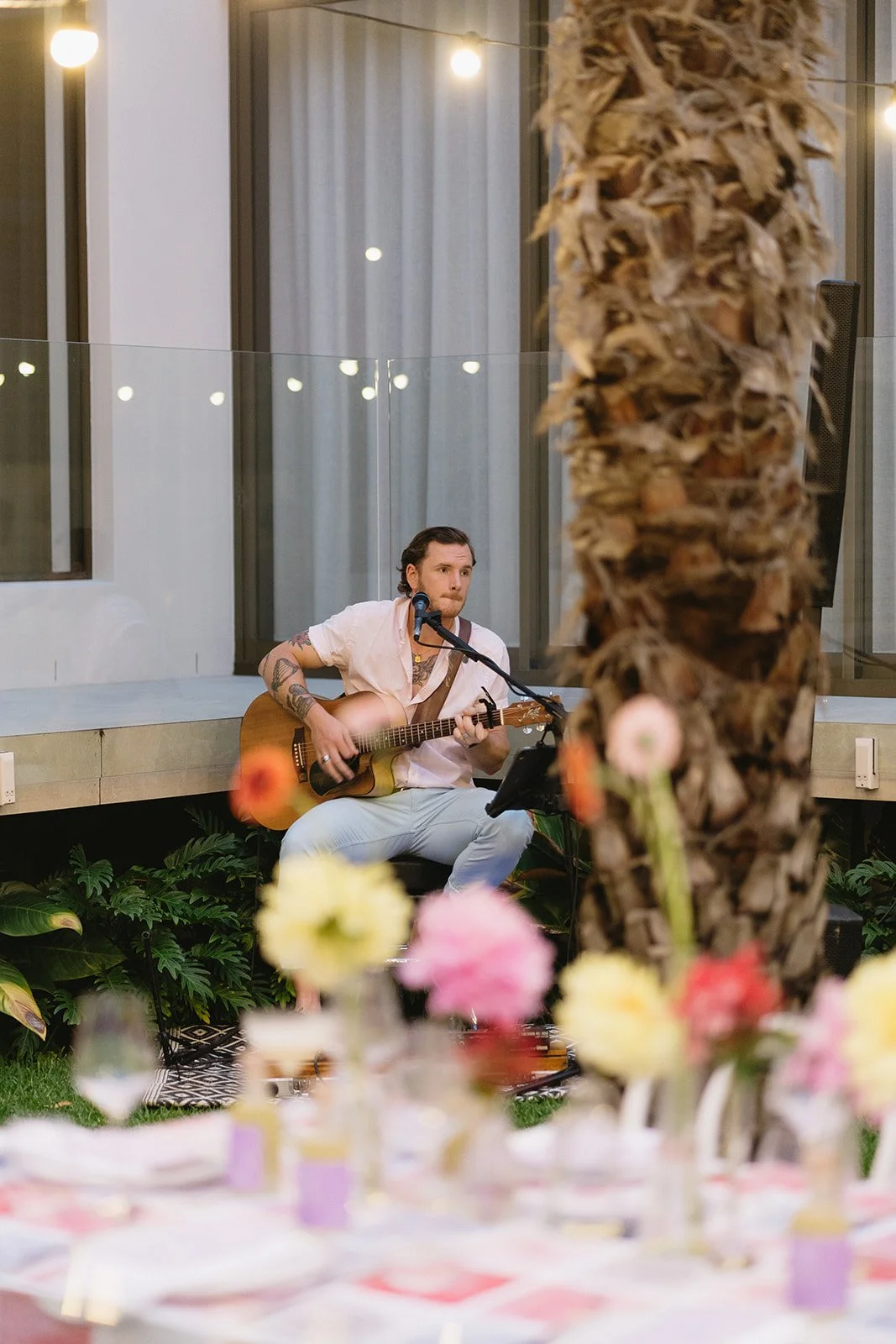 A man playing an acoustic guitar and singing into a microphone at an outdoor event, with a large palm tree and colorful flowers in the foreground.