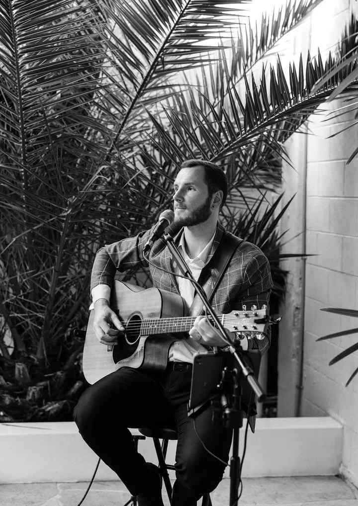 A man playing acoustic guitar and singing into a microphone outdoors, surrounded by palm leaves.