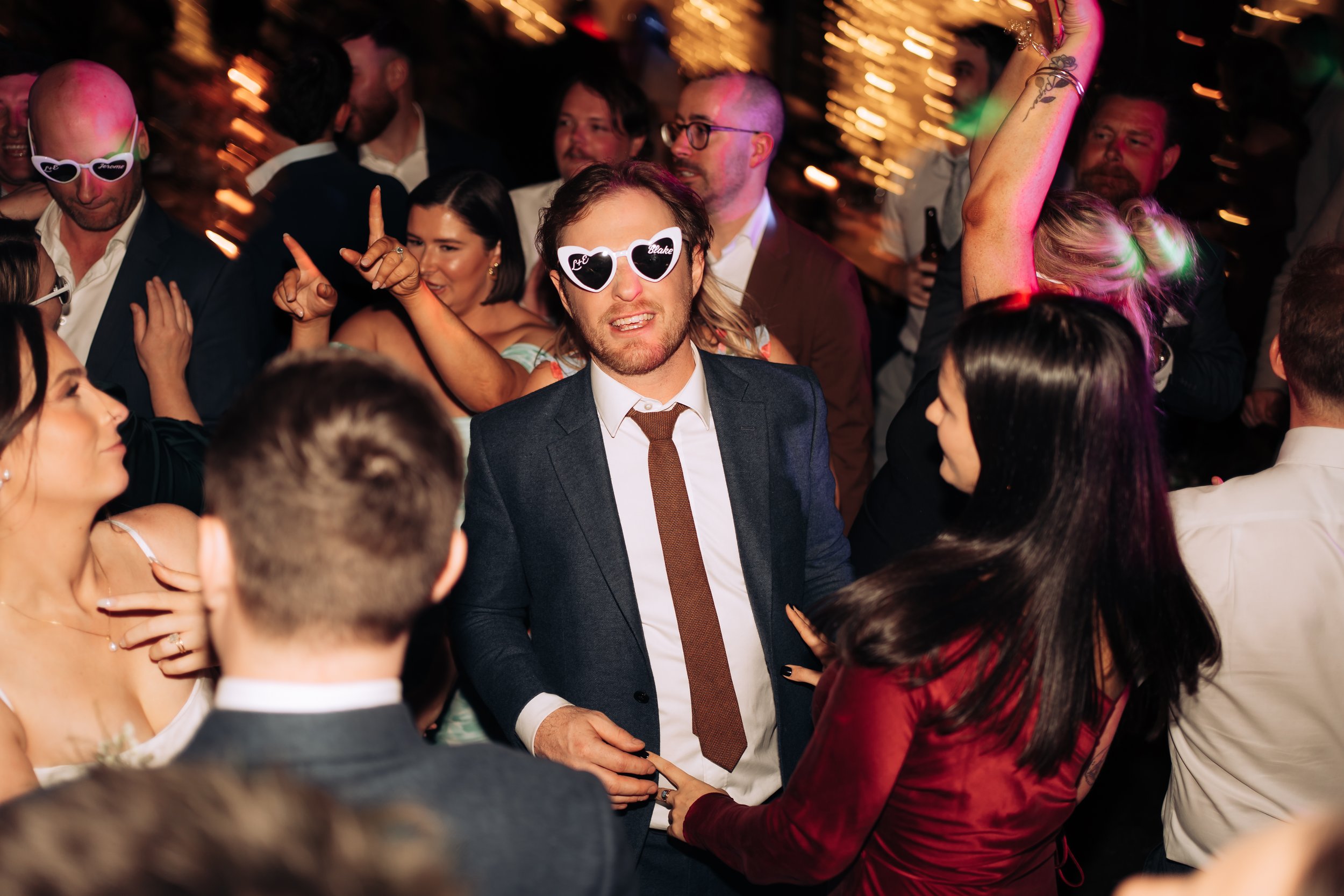 Crowd of people dancing and enjoying at a party, with some wearing sunglasses, in a lively atmosphere with colorful lights.