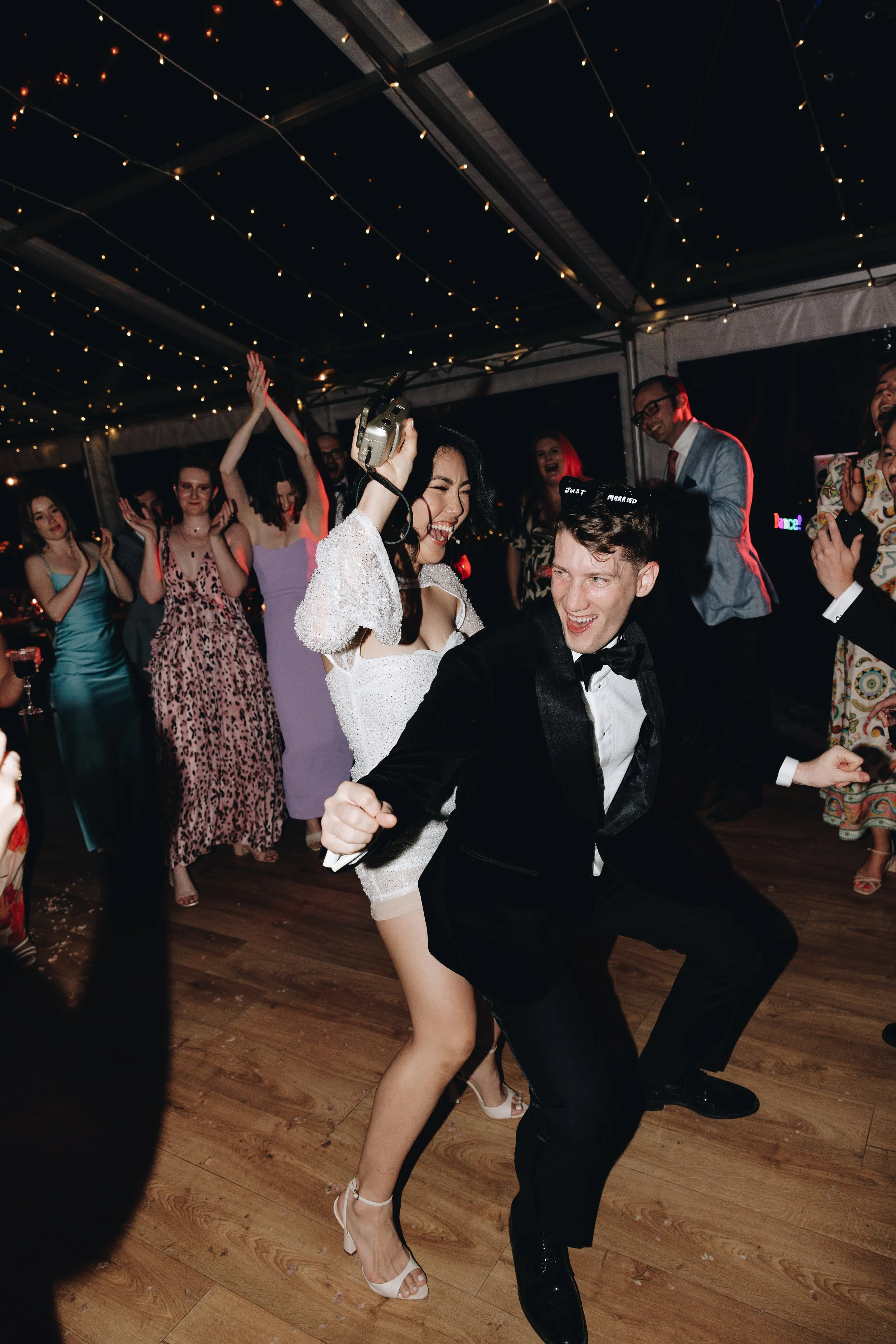 A couple dancing at a wedding reception with guests cheering around them, under ceiling string lights.