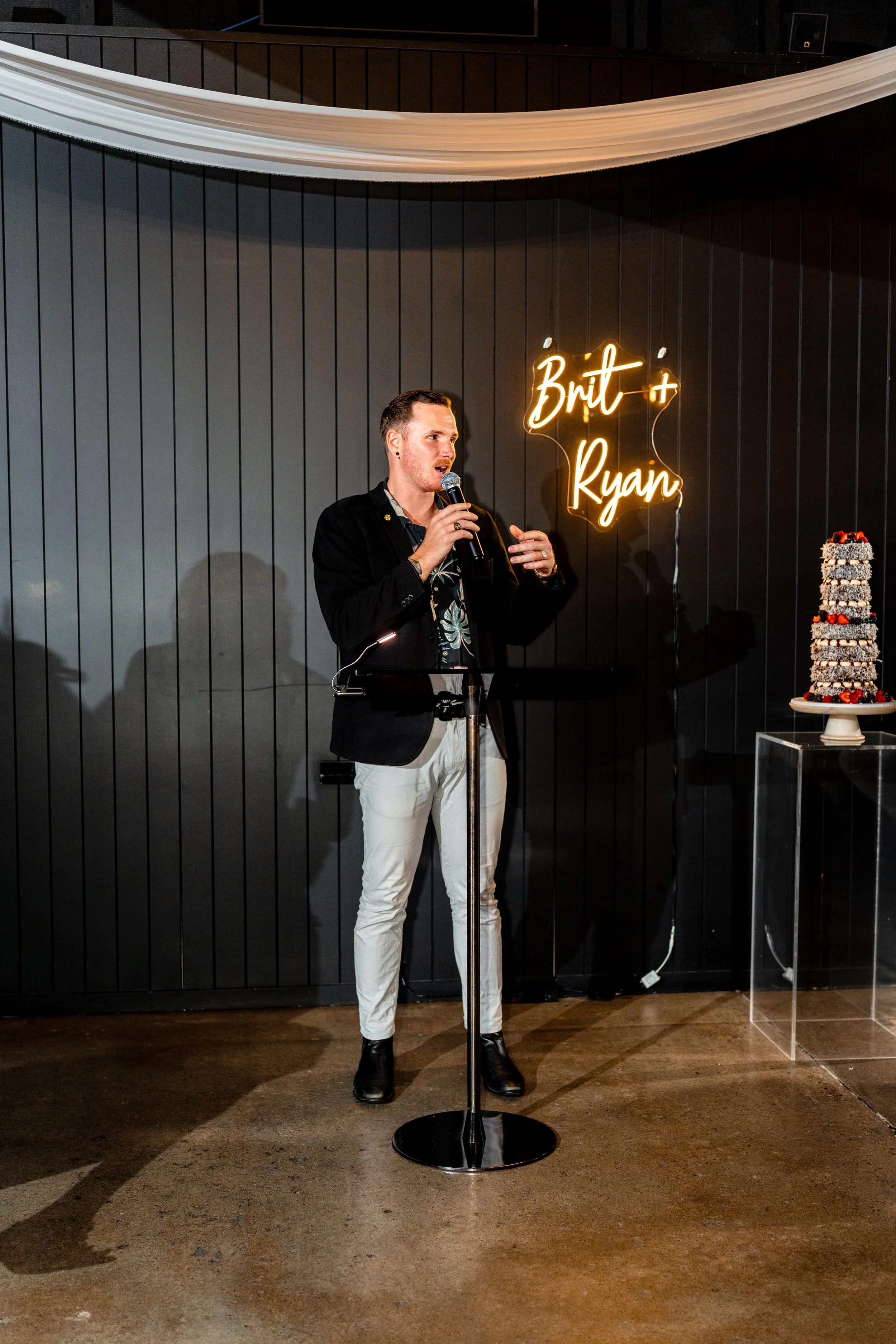 A man standing at a microphone in front of a dark wall, with a neon sign reading 'Britt + Ryan' behind him. To the right, there is a tall, multi-layered cake with chocolate and berries on a clear stand.