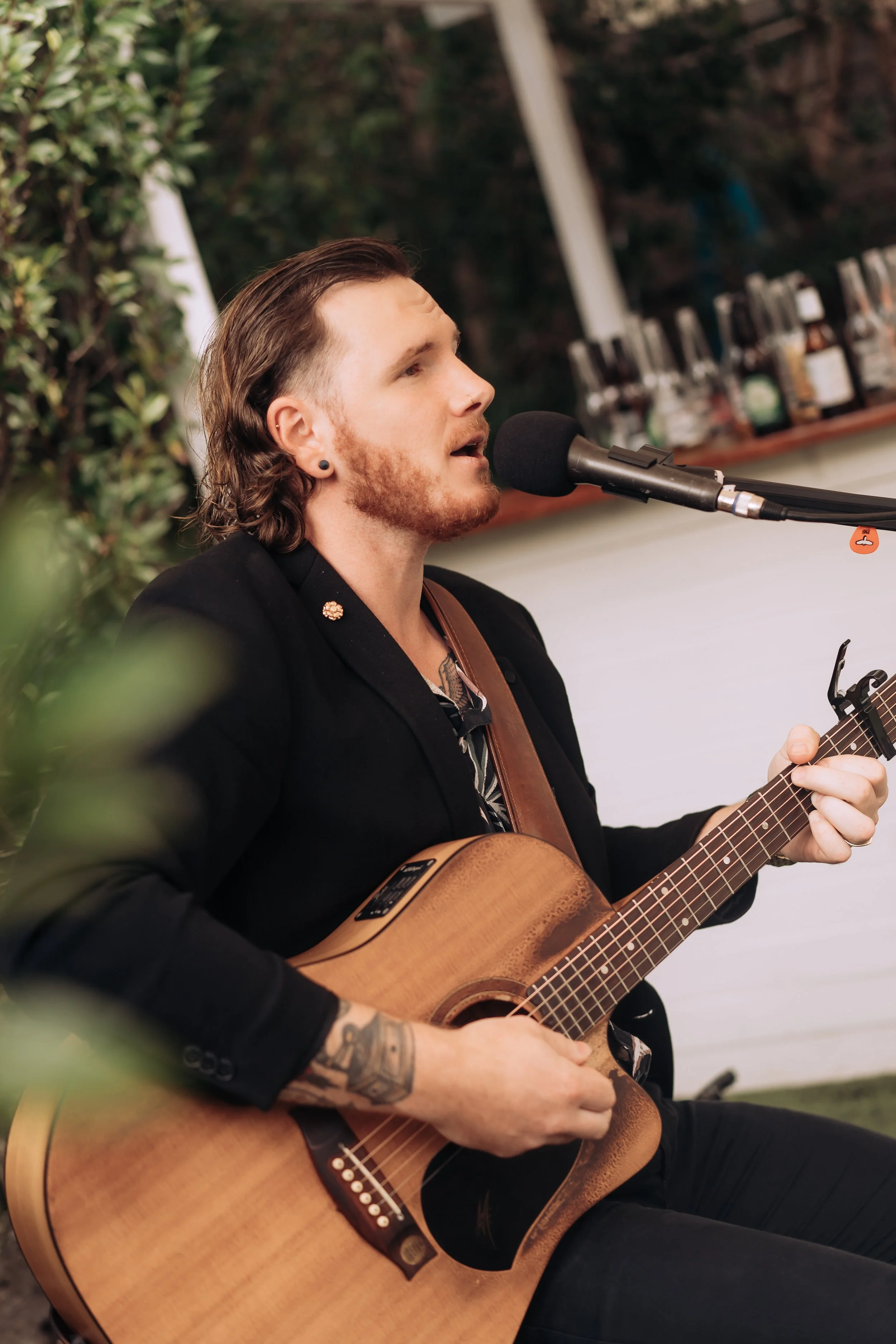 A man with long hair, a beard, and tattoos playing an acoustic guitar and singing into a microphone at an indoor event.