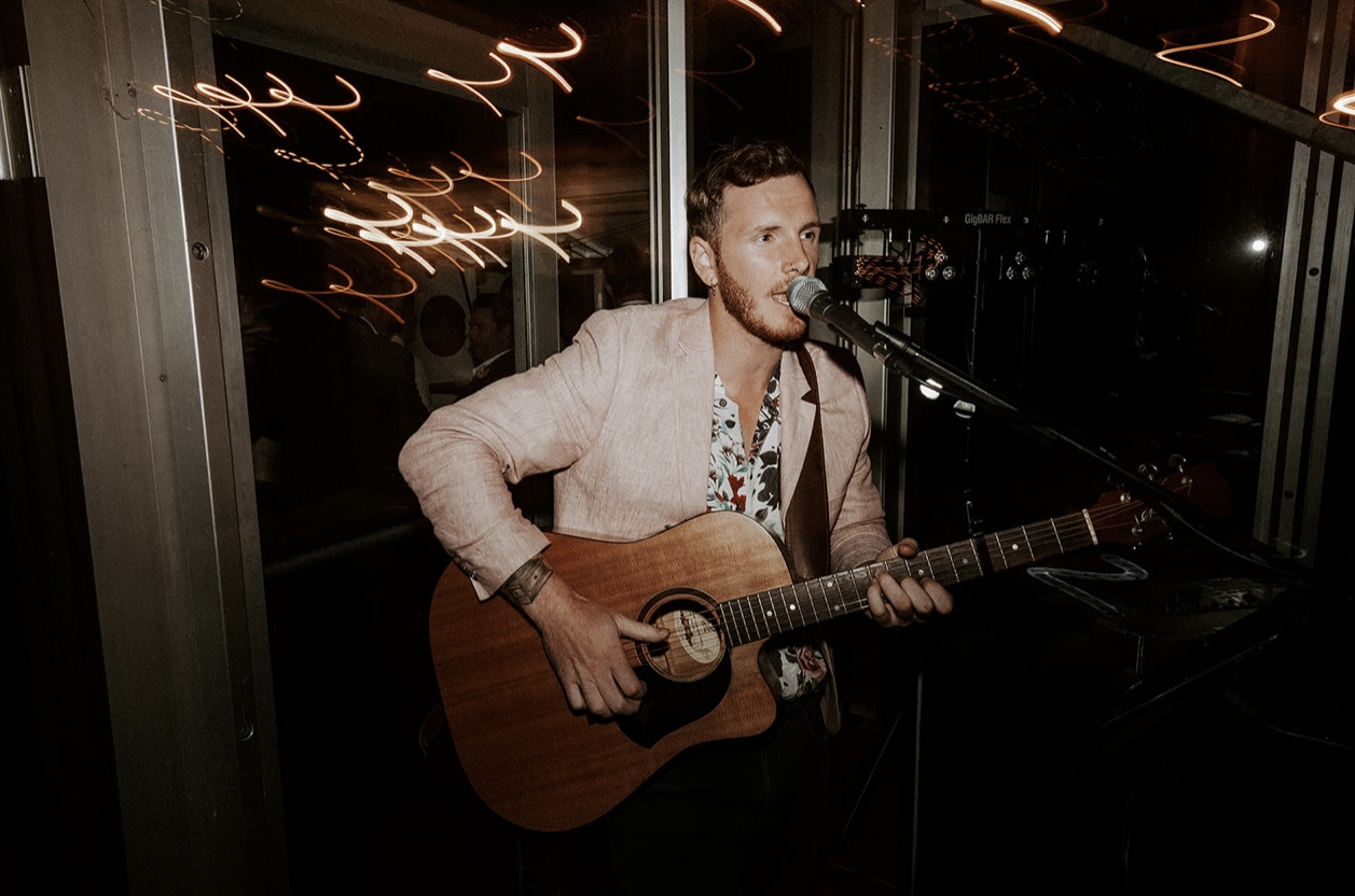 A man with short hair and a beard singing into a microphone while playing an acoustic guitar at a live music venue with warm, swirling light reflections in the background.