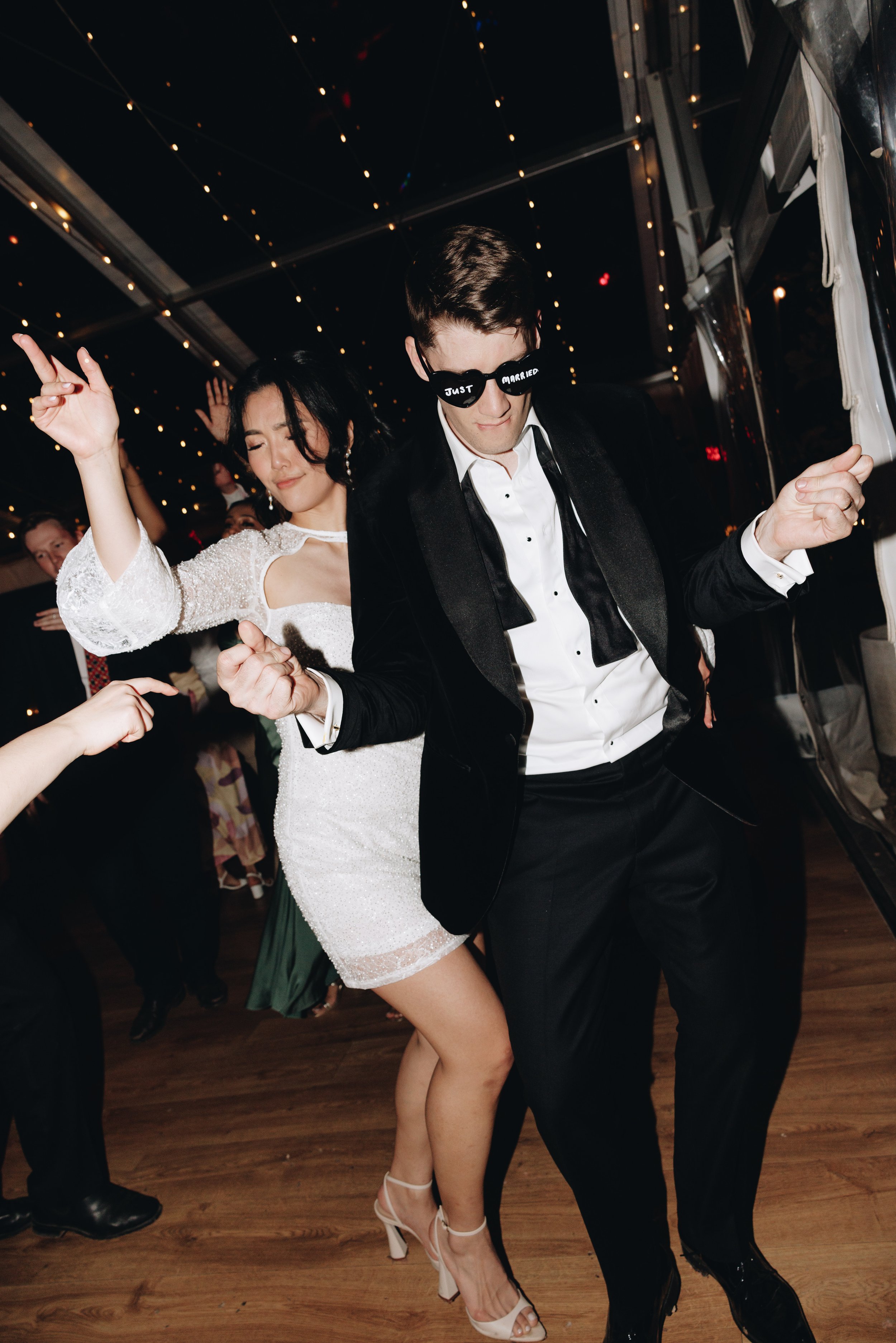 Couple dancing at a wedding reception, with the man wearing sunglasses that say "Just Married" and a tuxedo, and the woman wearing a white dress and high heels, surrounded by other guests on a wooden dance floor under string lights.