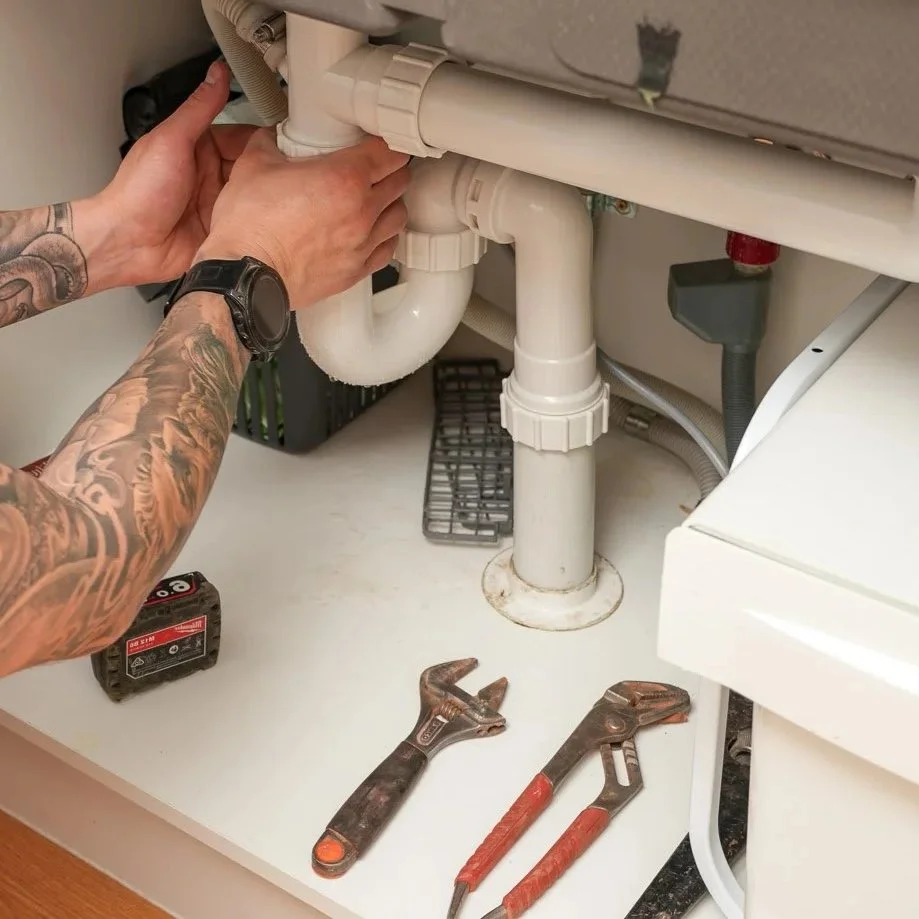 A plumber in a white shirt fixing plumbing under a kitchen sink, with tools including a wrench, pliers, and a drill on the counter.