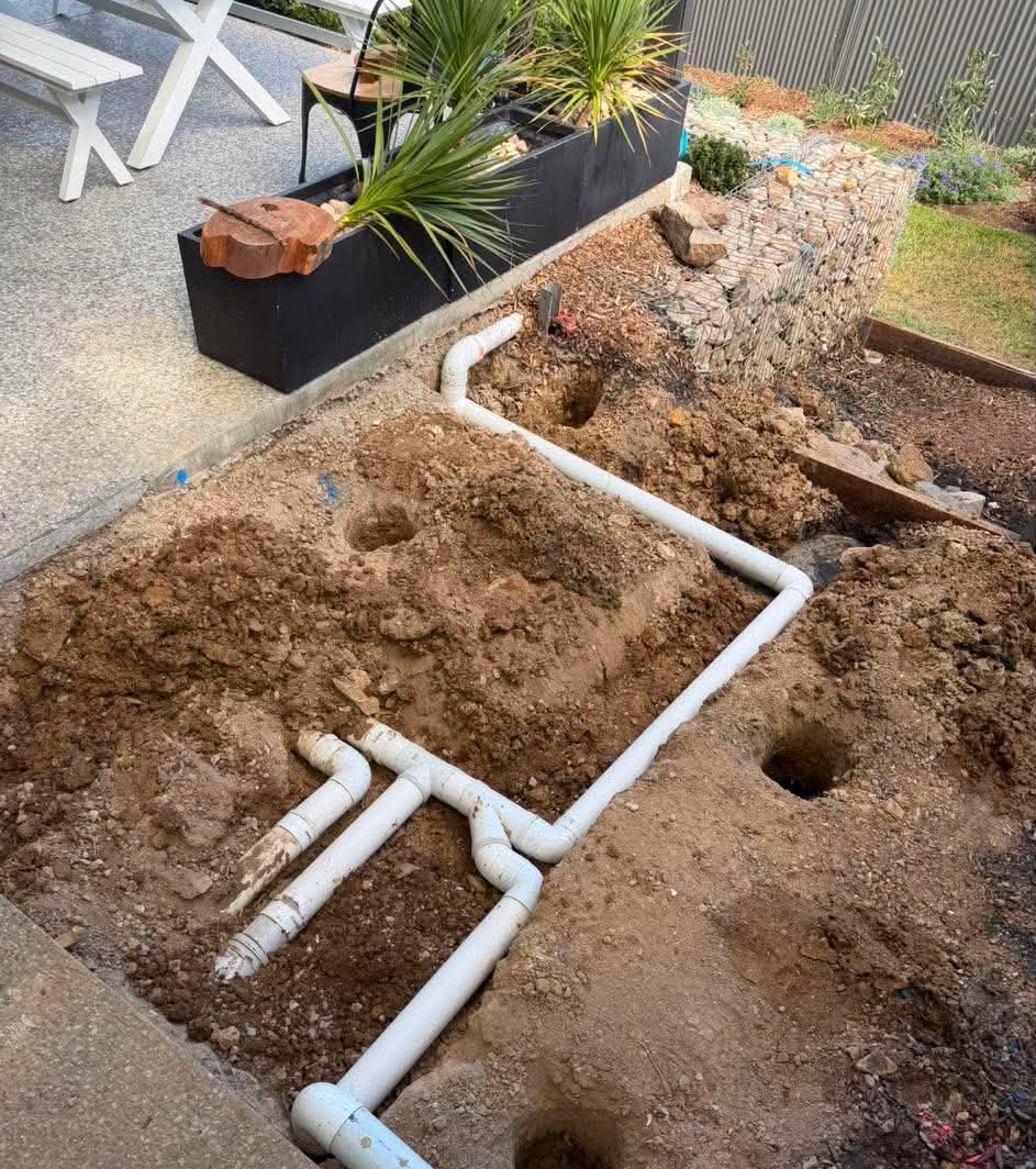 An outdoor excavation area showing a network of white PVC stormwater drainage pipes and dug post holes in clay soil, next to a concrete patio and a gabion retaining wall.