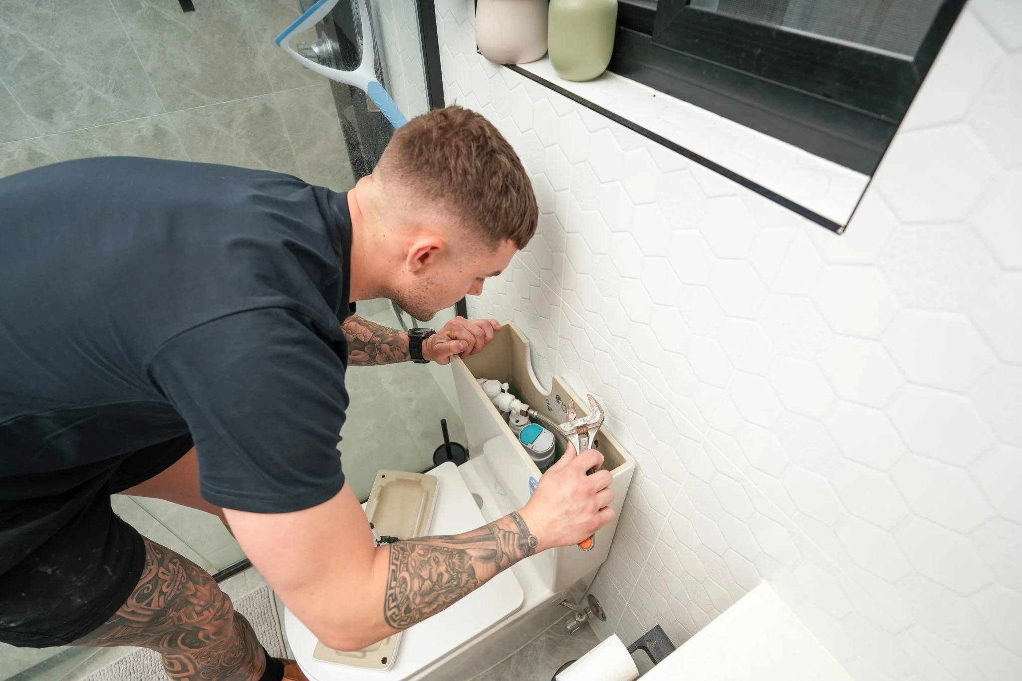 Emergency plumber using a shifting wrench to fix a leaking toilet cistern in a modern tiled bathroom.