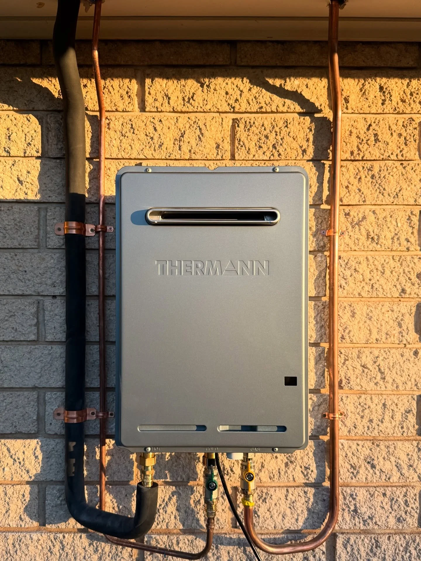 Close-up of a modern grey Thermann continuous flow gas hot water system installed against an outdoor brick wall.