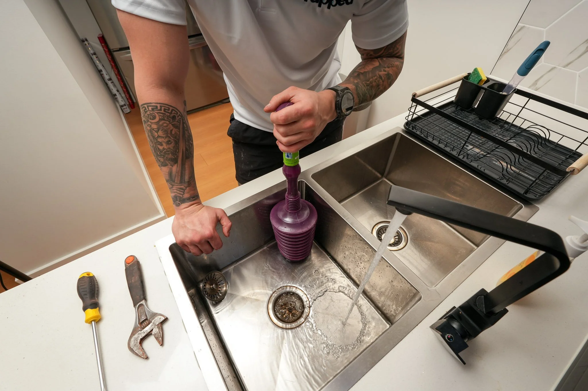 Overhead view of a plumber clearing a blocked double kitchen sink using a plunger, with tools laid out on the benchtop.