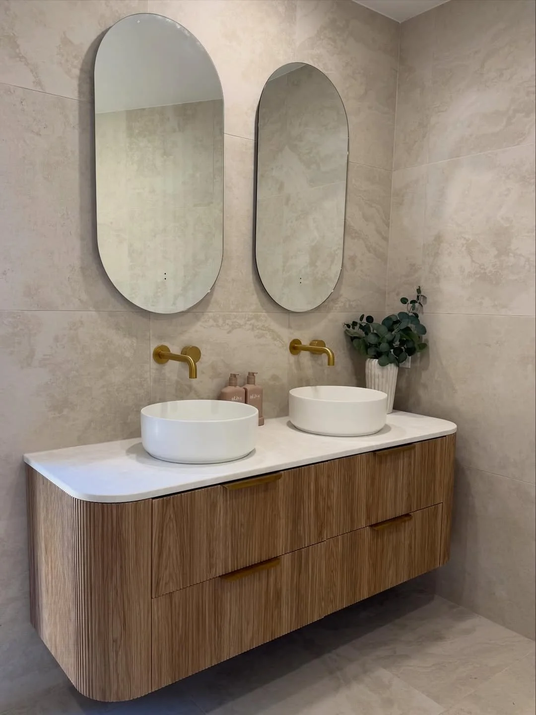 Modern luxury bathroom renovation featuring dual white vessel basins on a timber vanity with brushed gold tapware and round mirrors.