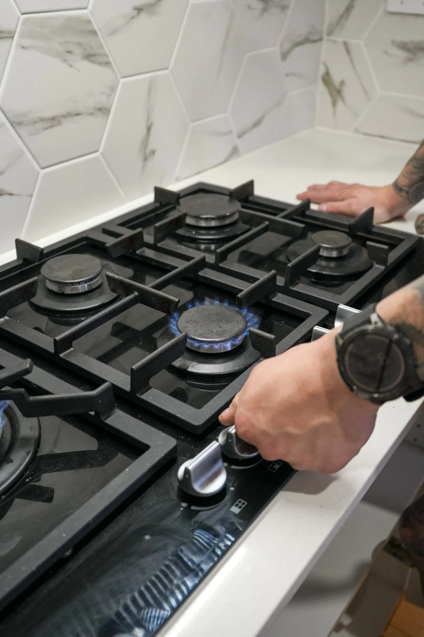 A person adjusting the control knob on a black gas stove with four burners, with one burner lit and blue flames visible. The stove is installed on a white countertop with a white and gray checkered tile backsplash.