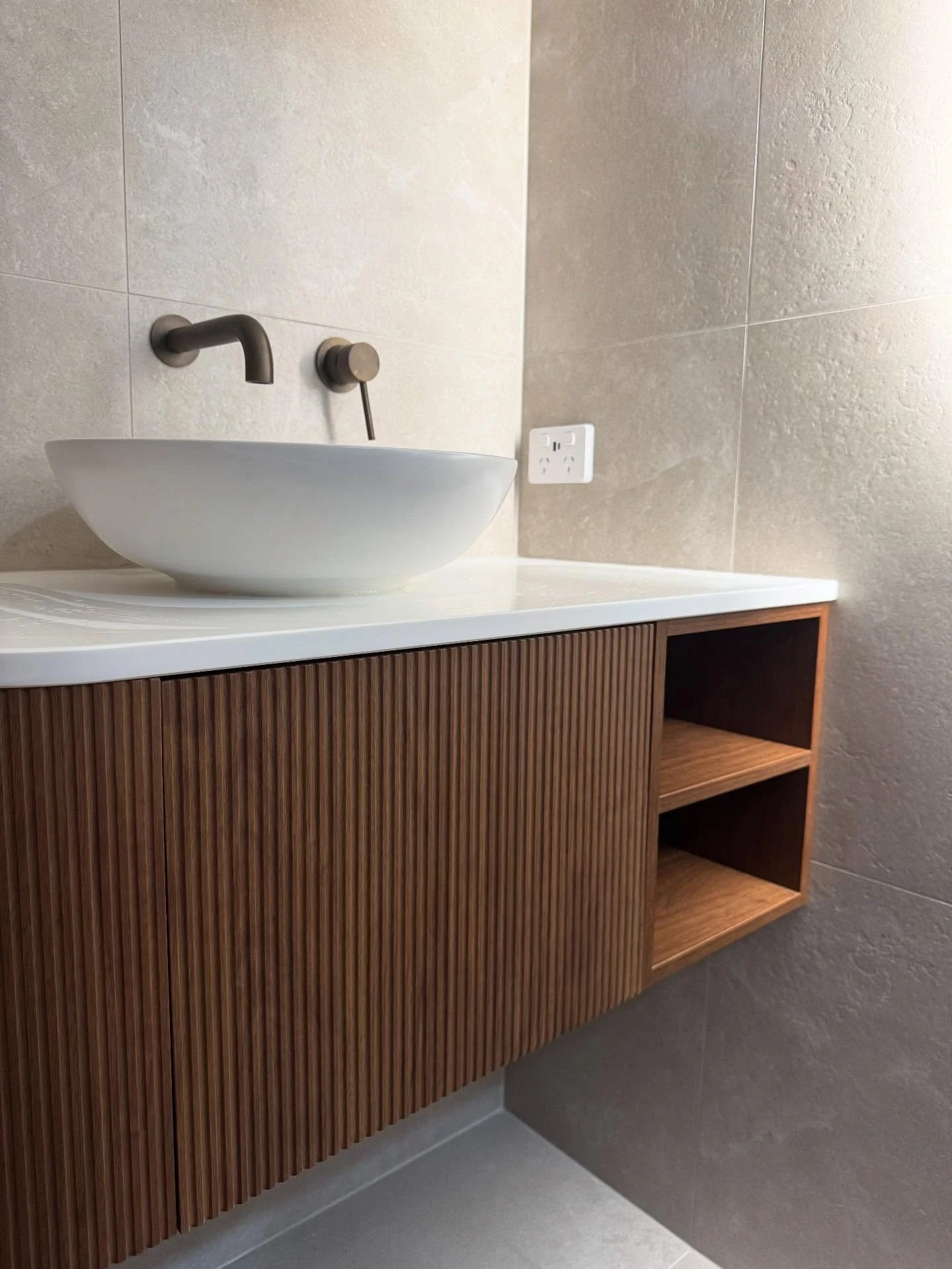 Modern bathroom sink with vessel basin, wall-mounted faucet, and wooden vanity with open shelves, beige tiled walls, and electrical outlet.