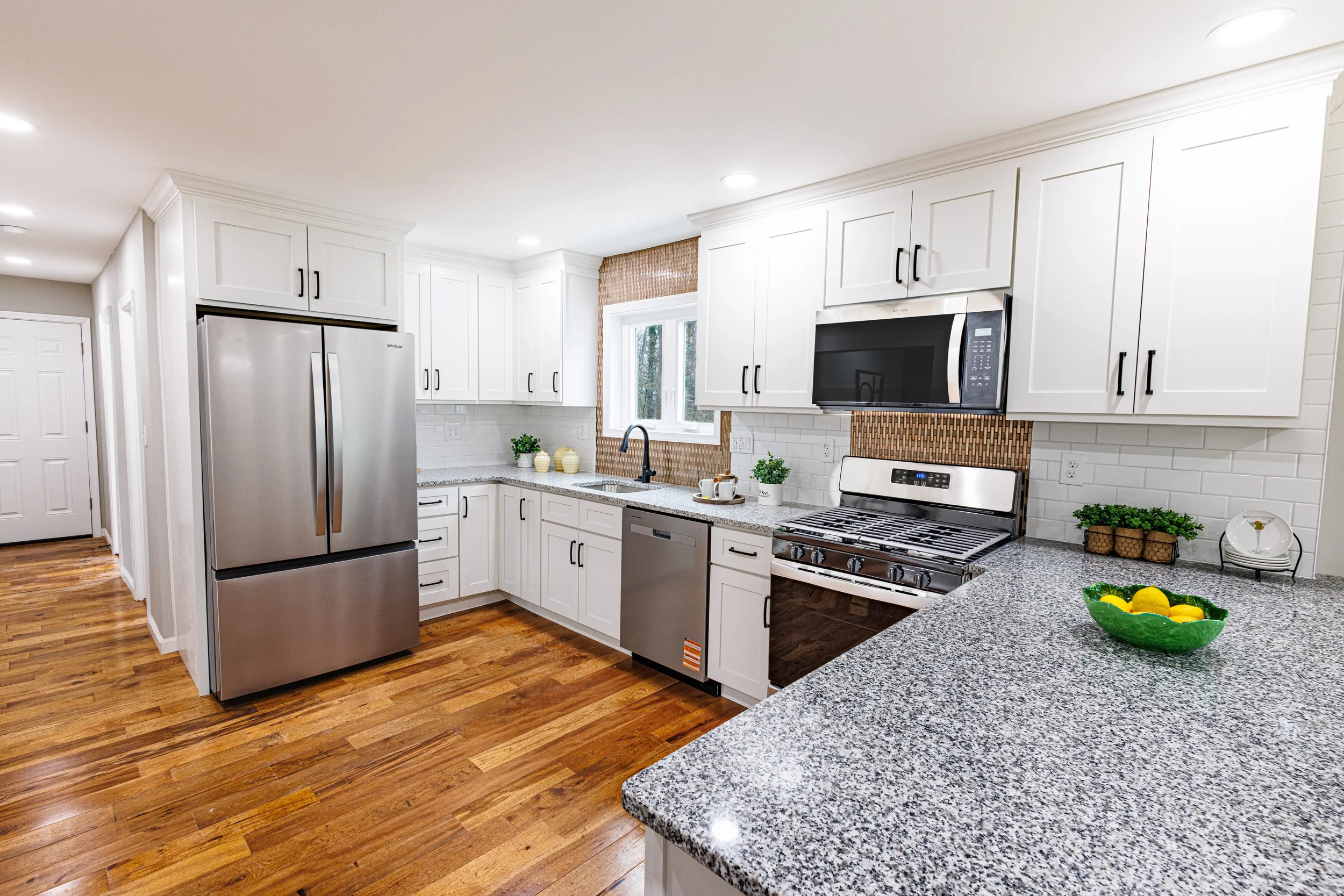 A modern kitchen with white cabinets, stainless steel refrigerator and oven, granite countertops, hardwood floors, and a window with a view outside. Decor includes small potted plants and bowls of lemons.