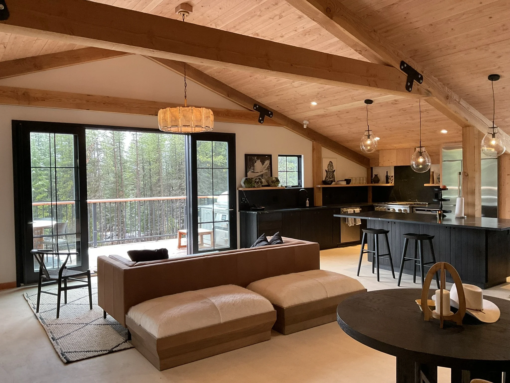 Living room with wood-paneled vaulted ceiling, large sliding glass doors leading to a deck with outdoor furniture, a beige sofa, a patterned rug, and multiple hanging pendant lights.