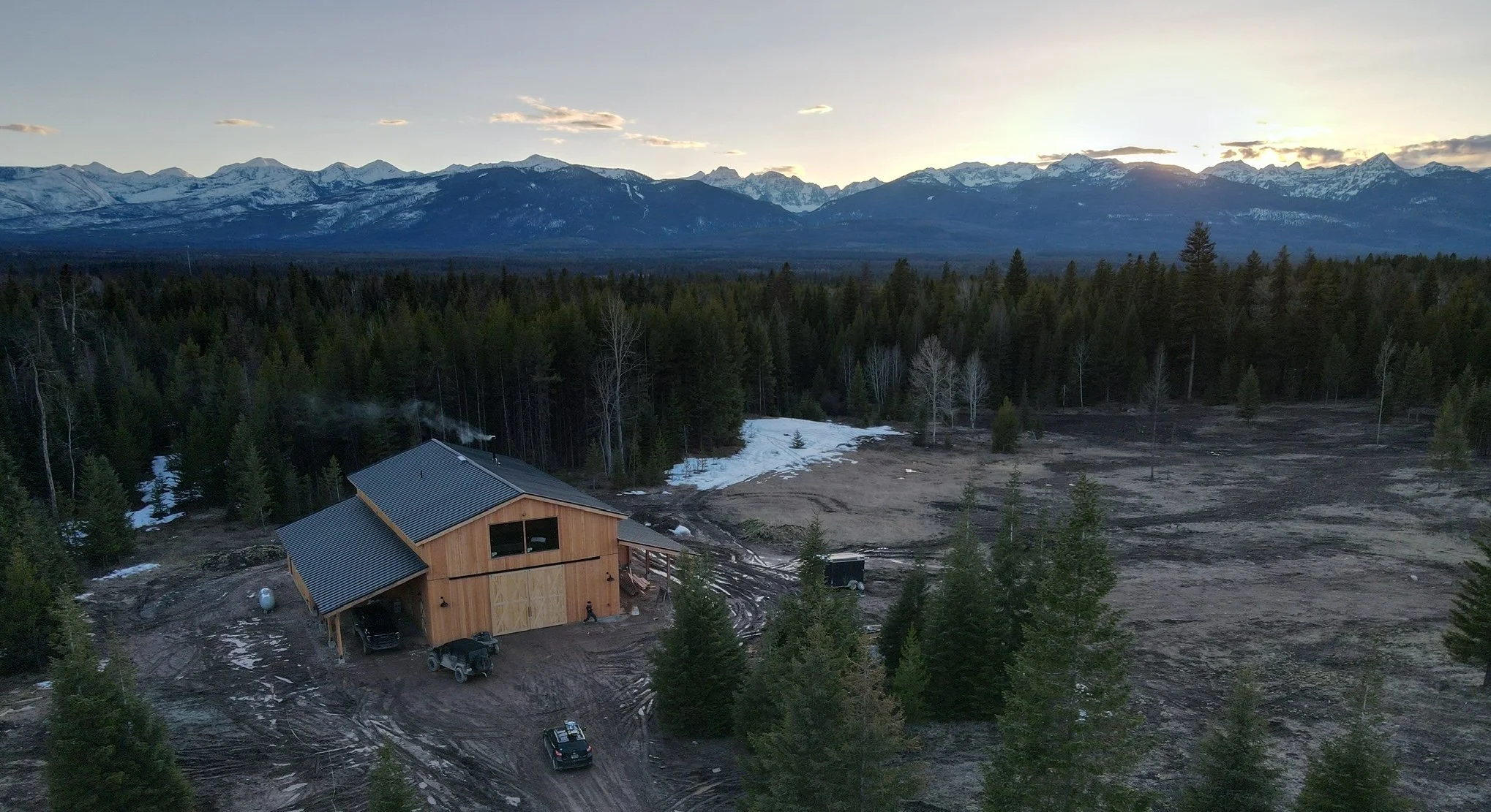 A landscape scene showing a house in the foreground with construction activity and vehicles nearby, a dense forest, and snow-capped mountains in the background under a partly cloudy sky.