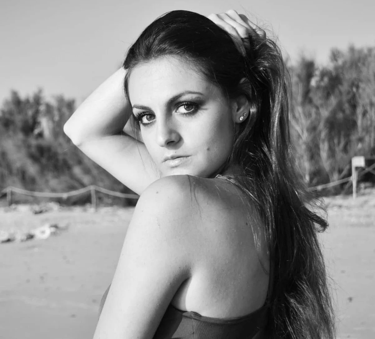 Black and white photograph of a woman with long dark hair, looking at the camera, with her hand on her head, outdoors with trees and a beach in the background.
