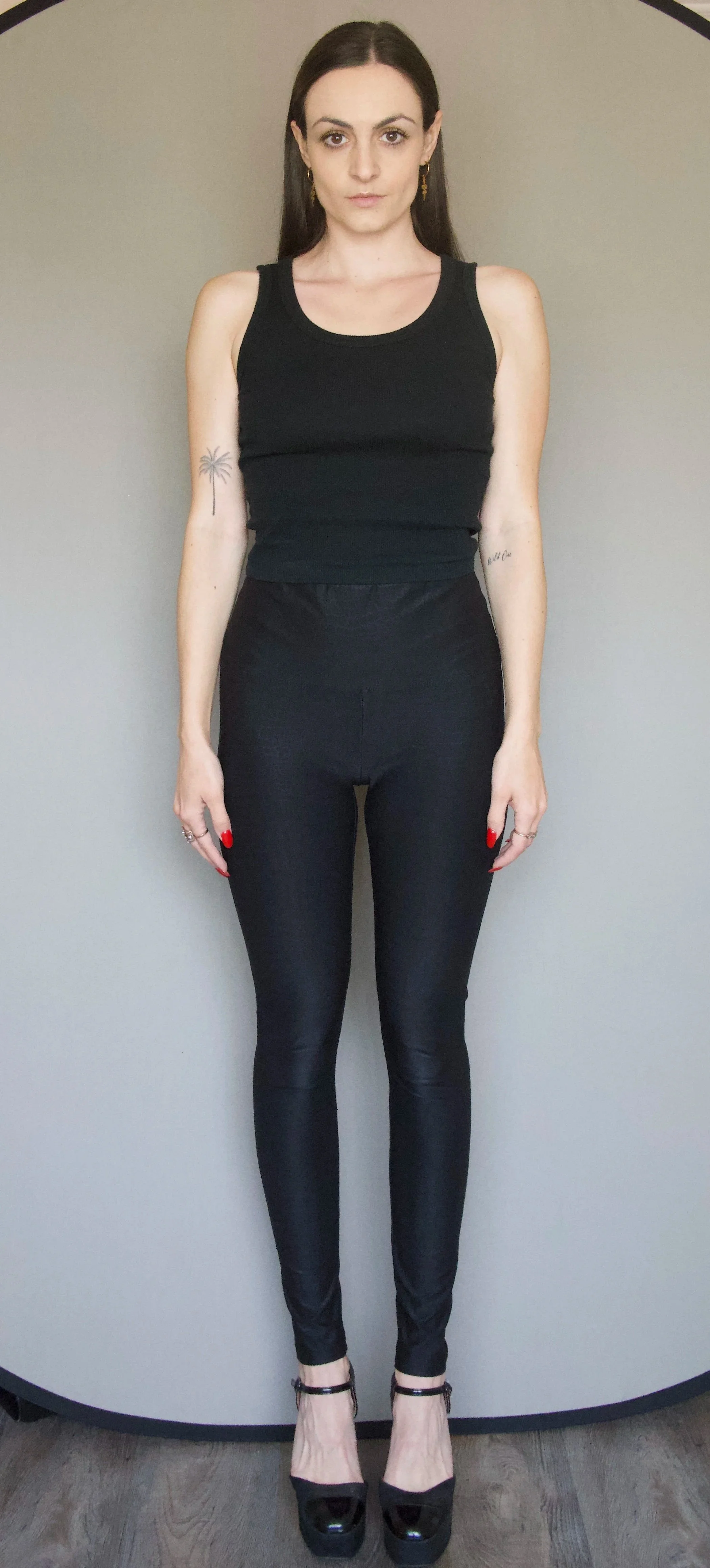 A woman with dark hair pulled back, wearing a black tank top, black pants, and black high heels, standing against a plain wall.