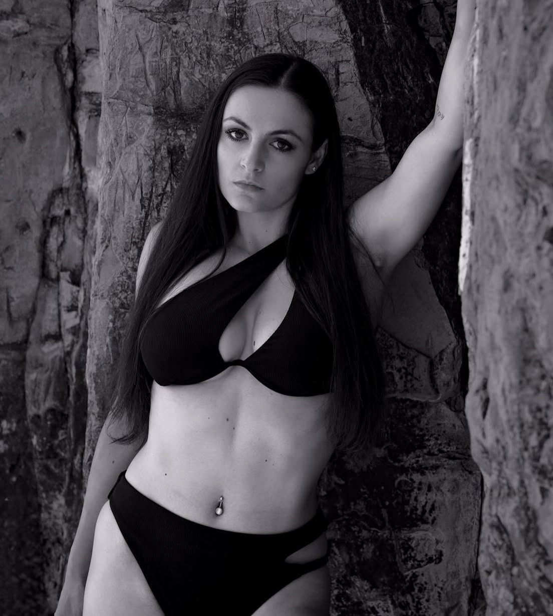A young woman with long dark hair and light skin posing against rock formations, wearing a black bikini top and matching bottoms, with a piercing on her navel, looking directly at the camera.