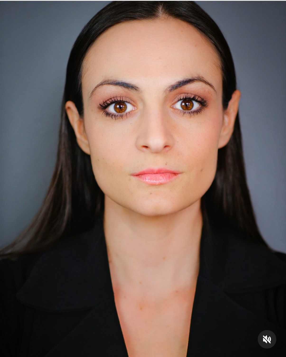 Close-up portrait of a woman with long dark hair, brown eyes, and natural makeup, wearing a black blazer, against a plain gray background.