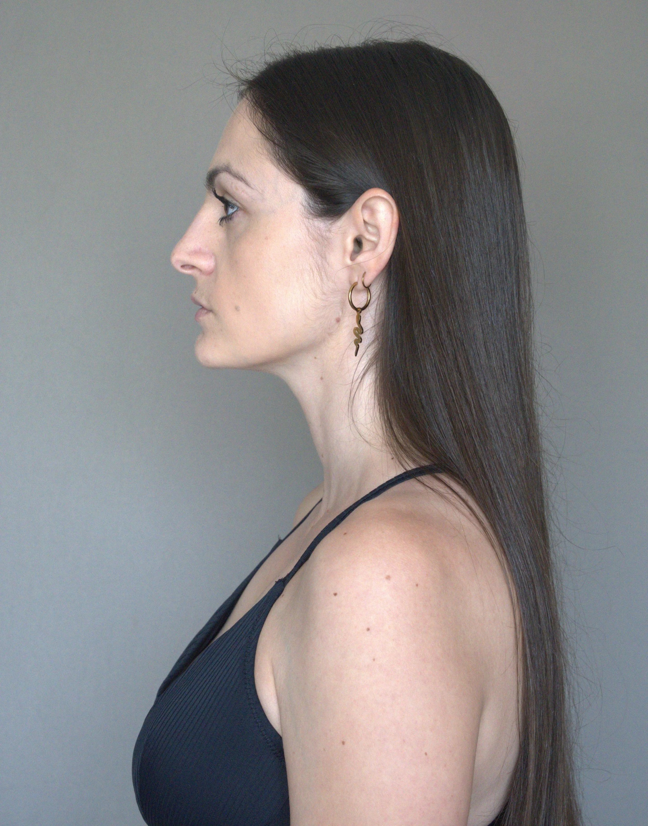 Profile of a woman with long dark hair, wearing a black tank top and gold earrings, against a plain gray background.