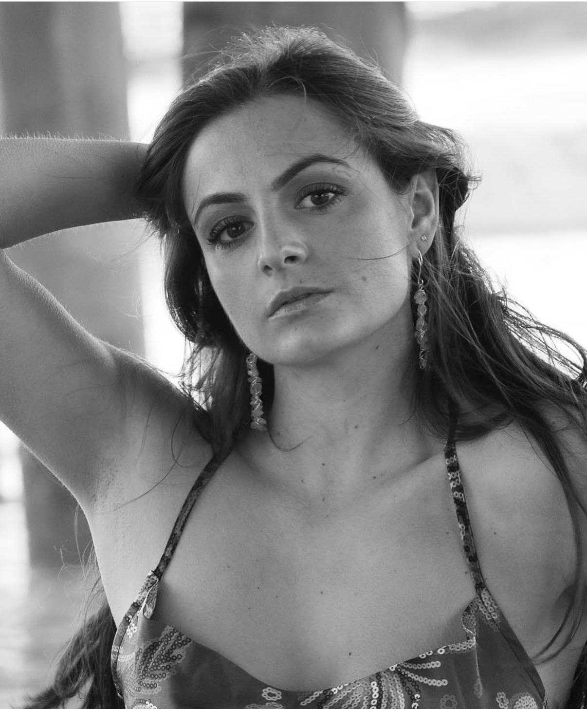 Black-and-white portrait of a woman with long hair, wearing earrings and a patterned top, looking directly at the camera with one arm raised behind her head.