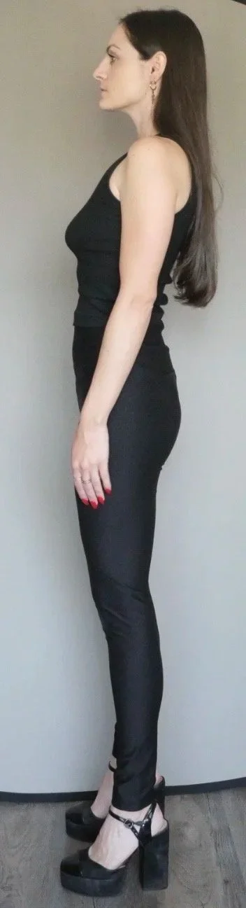 Side view of a woman with long dark hair wearing a black sleeveless top, black leather pants, and black high heels, standing against a plain wall.