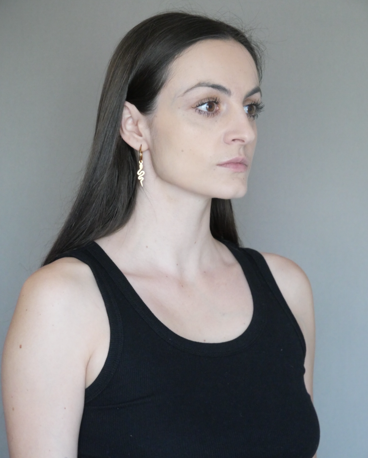 Profile of a woman with long dark hair, wearing a black tank top and gold earrings, against a gray background.