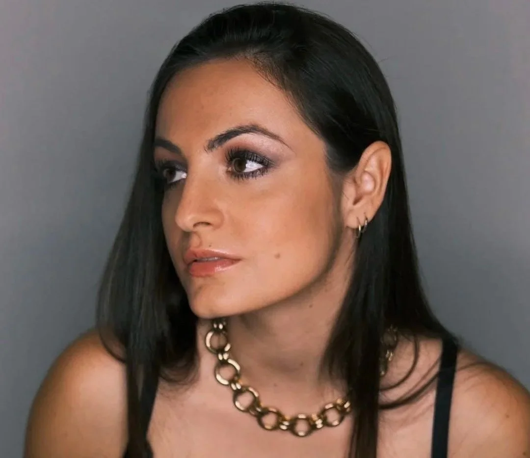 Portrait of a woman with long dark hair, gold jewelry, and makeup, against a gray background.