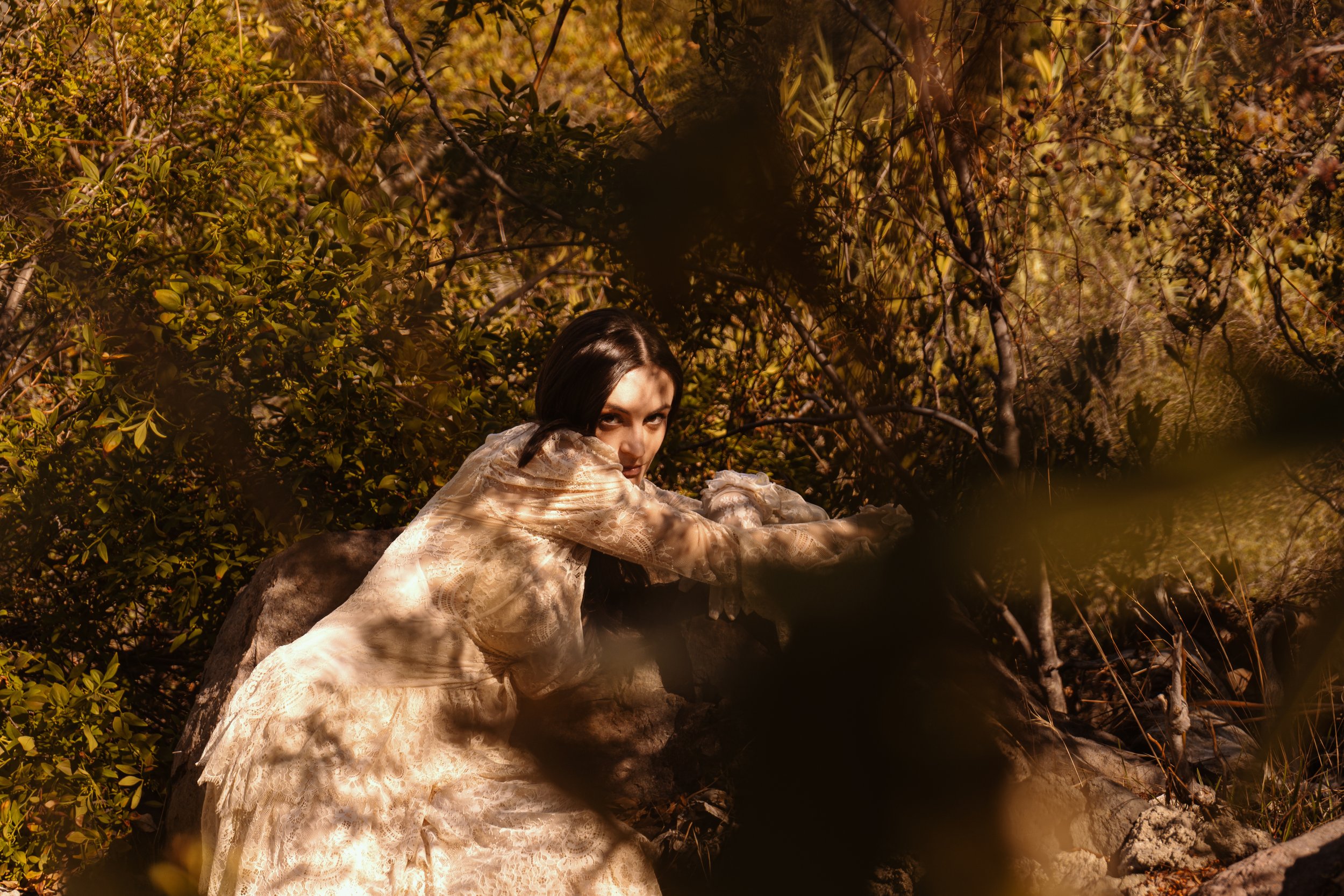 A woman with dark hair in a long, cream-colored lace dress crouches among dense foliage and rocks. She looks directly at the camera with a serious expression, blending into the natural surroundings.