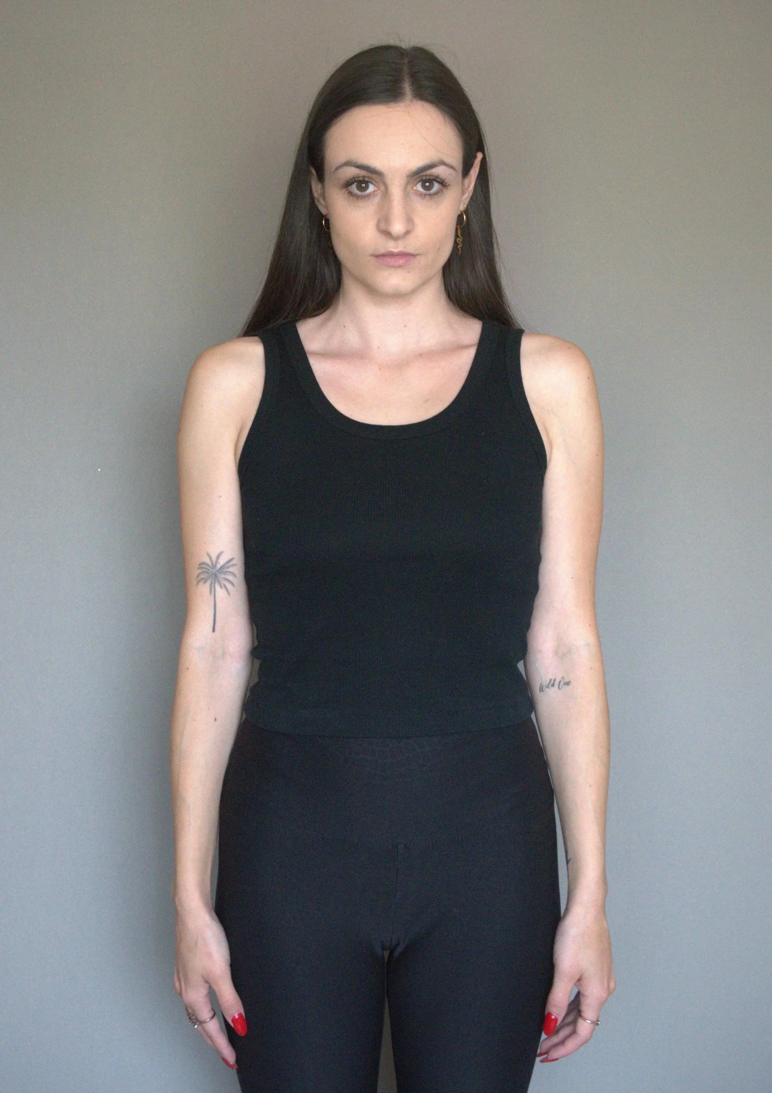 Young woman with long dark hair, wearing a black sleeveless top and black pants, standing against a gray background, with tattoos on her left arm and right wrist.