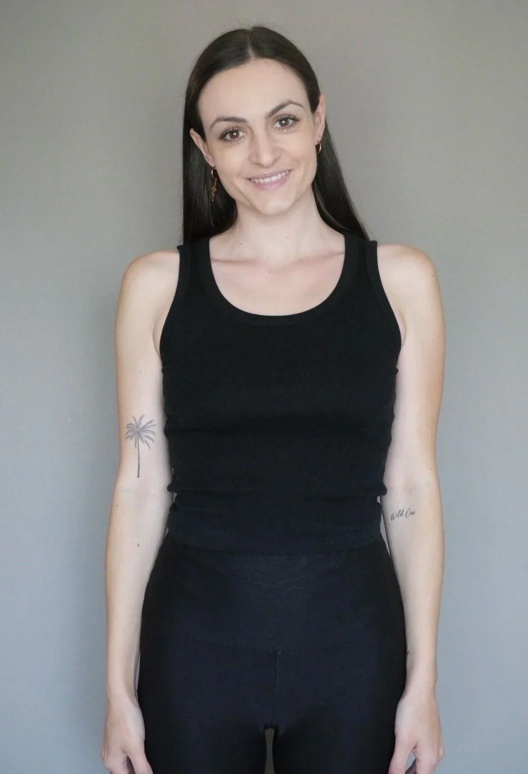 A young woman with long dark hair smiling, wearing a black tank top and black pants, standing against a plain gray background.