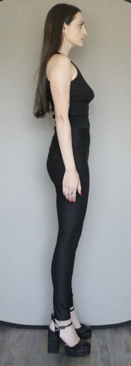 Side profile of a woman with long dark hair wearing a black sleeveless top, black tight pants, and black high heel shoes, standing against a plain gray wall.