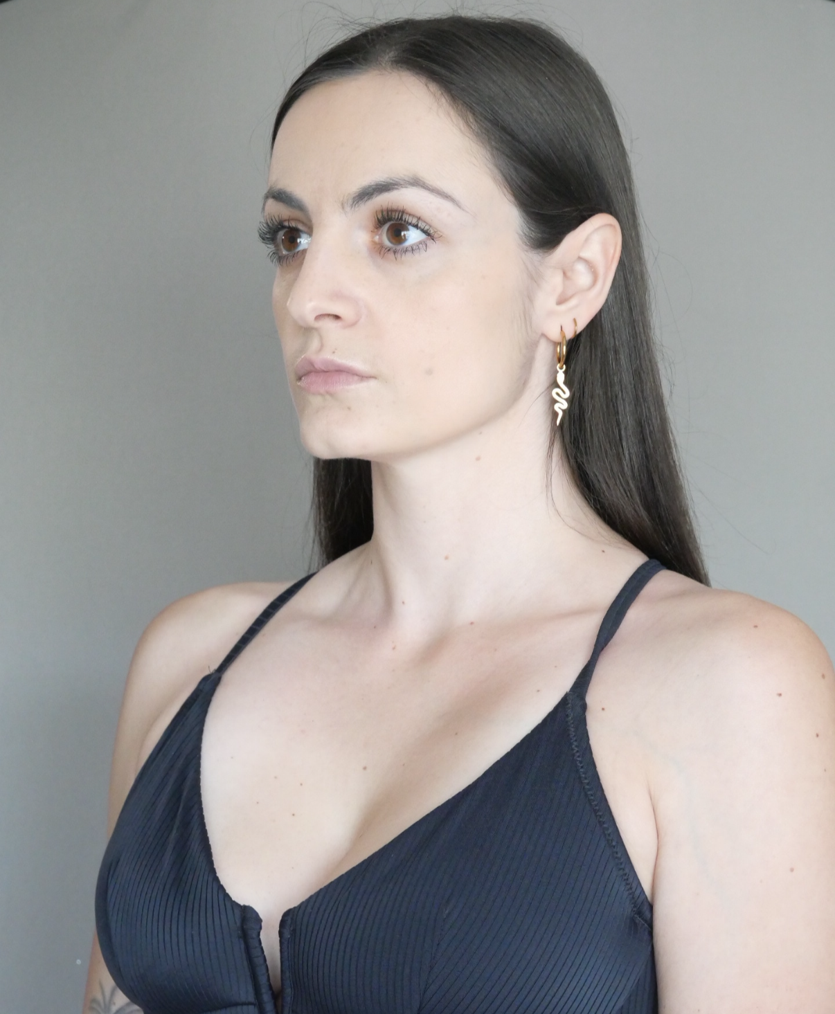 A woman with long dark hair, wearing a black tank top and gold snake-shaped earrings, looking slightly to the side against a neutral background.