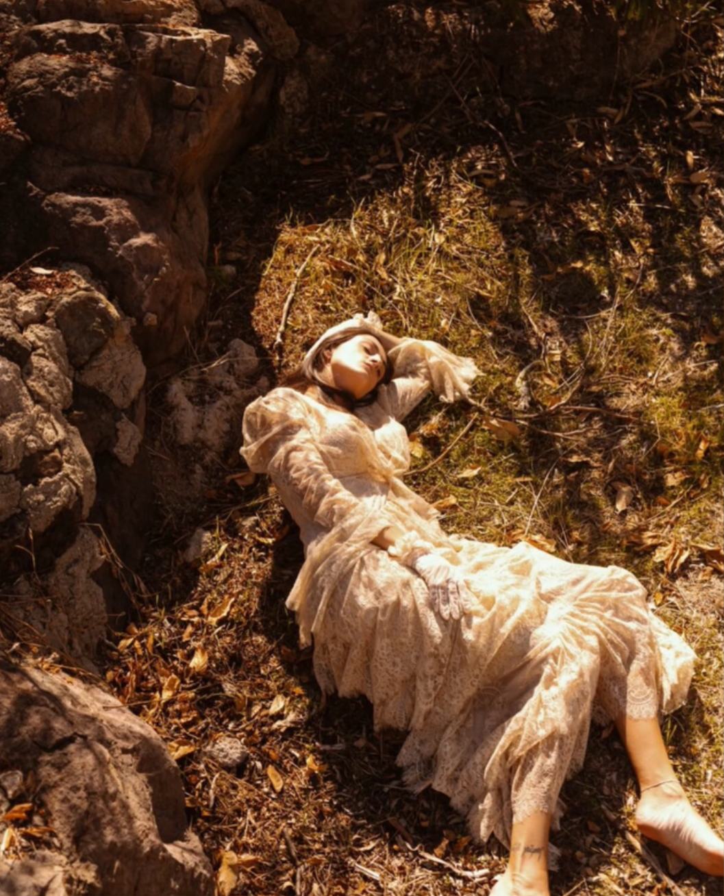 A woman dressed in vintage lace clothing lying on the ground amidst dry leaves and dirt, her eyes closed, appearing to be sleeping or resting.