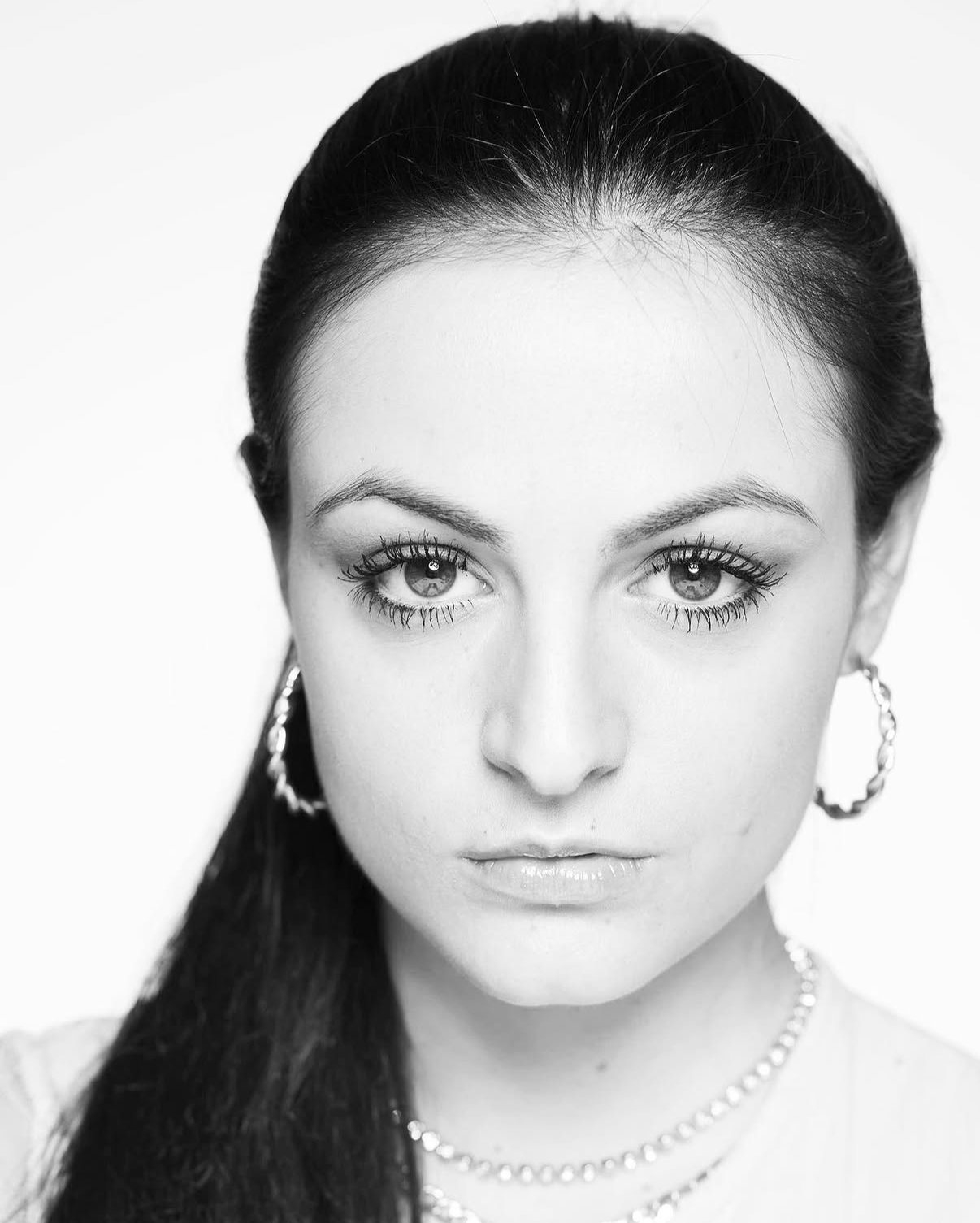 Close-up black and white portrait of a woman with dark hair, hoop earrings, and layered necklaces, looking directly at the camera.