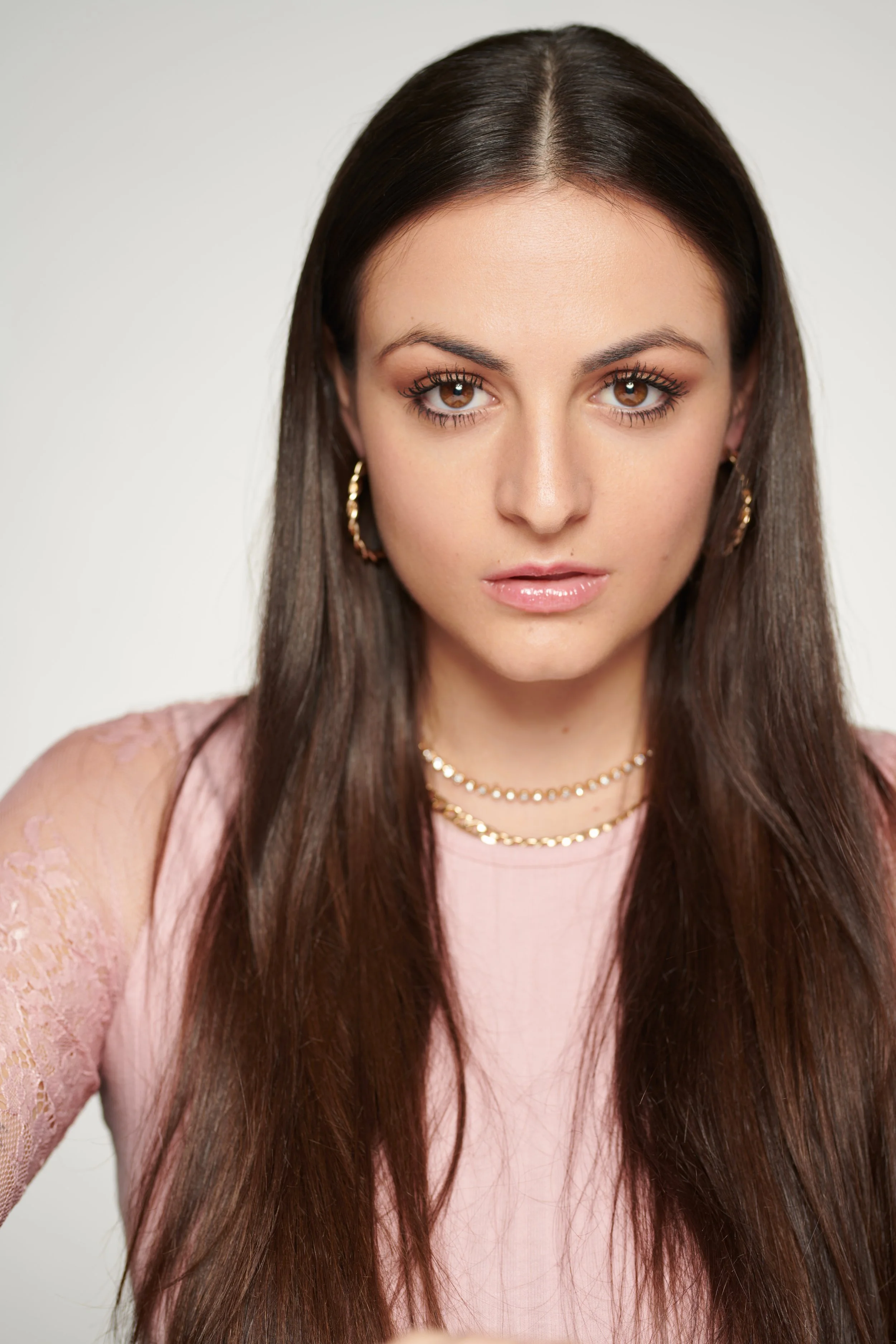 Close-up of a woman with long dark brown hair, brown eyes, and wearing pink lipstick, gold hoop earrings, and layered necklaces, looking directly at the camera against a plain light background.