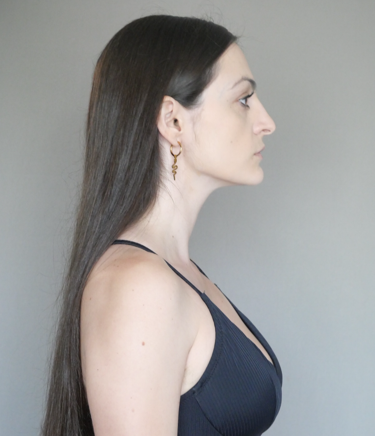 Profile side view of a woman with long dark hair, wearing a black tank top and a gold snake-shaped earring, against a plain gray background.
