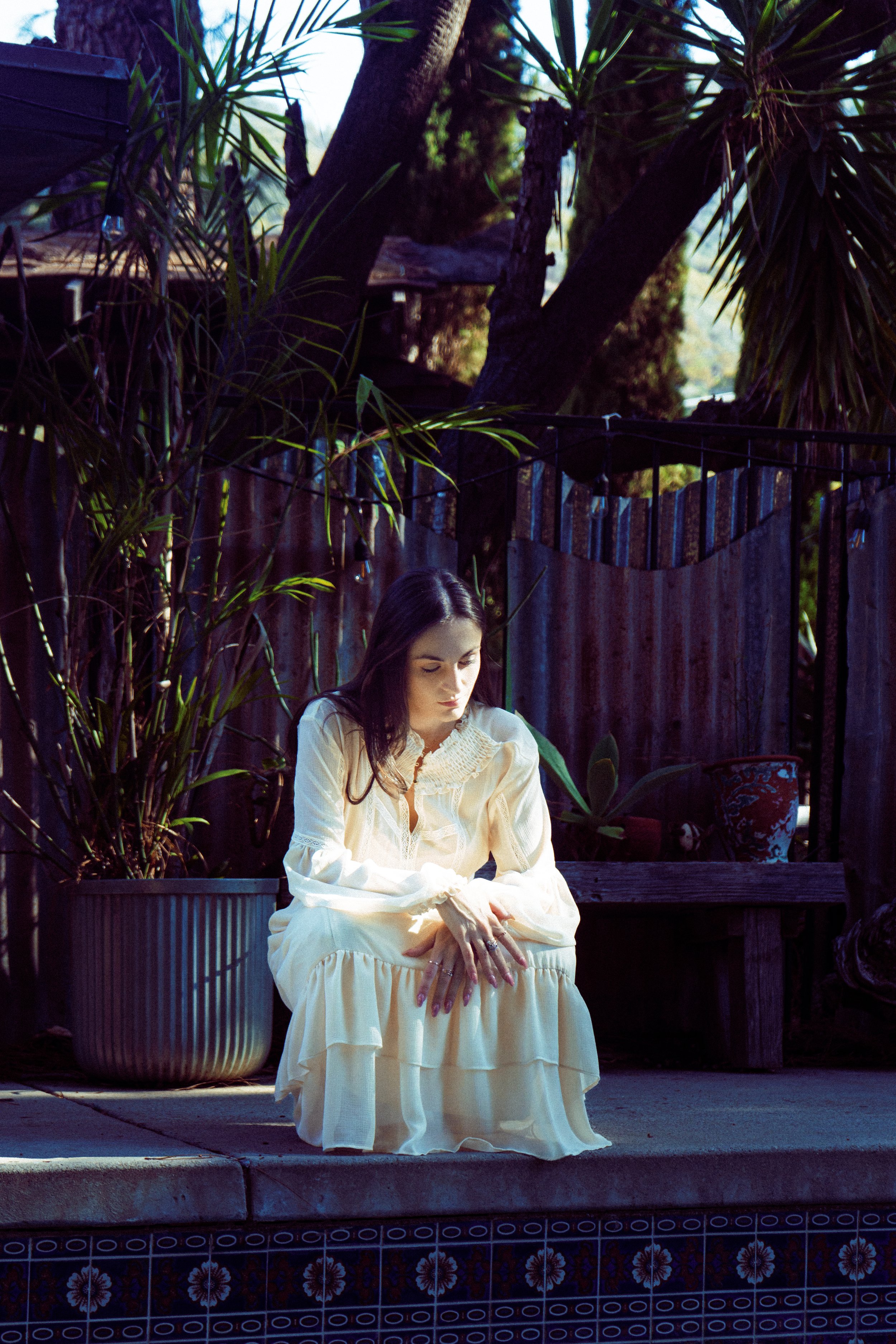 A woman in a cream-colored dress sitting on the edge of a swimming pool, surrounded by plants and a wooden fence.