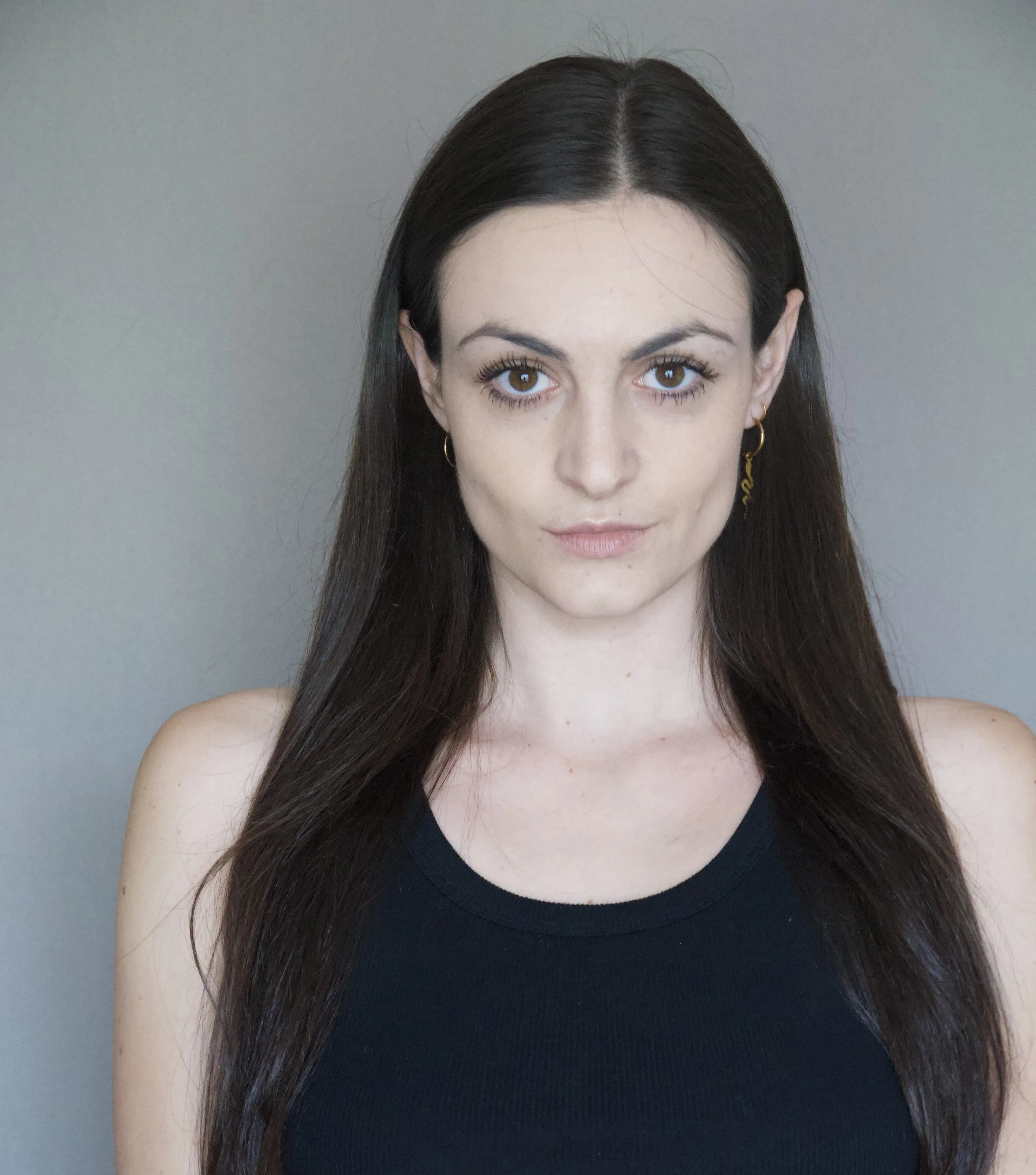 A young woman with long dark hair and brown eyes, wearing a black sleeveless top, standing against a plain gray background.