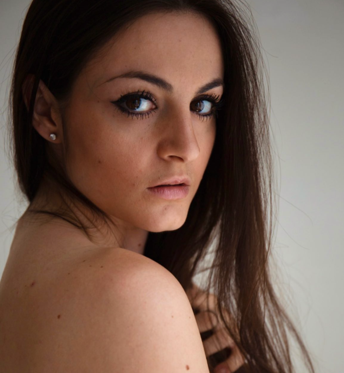 Close-up of a woman with dark brown hair, makeup with winged eyeliner, and earrings, looking over her shoulder.