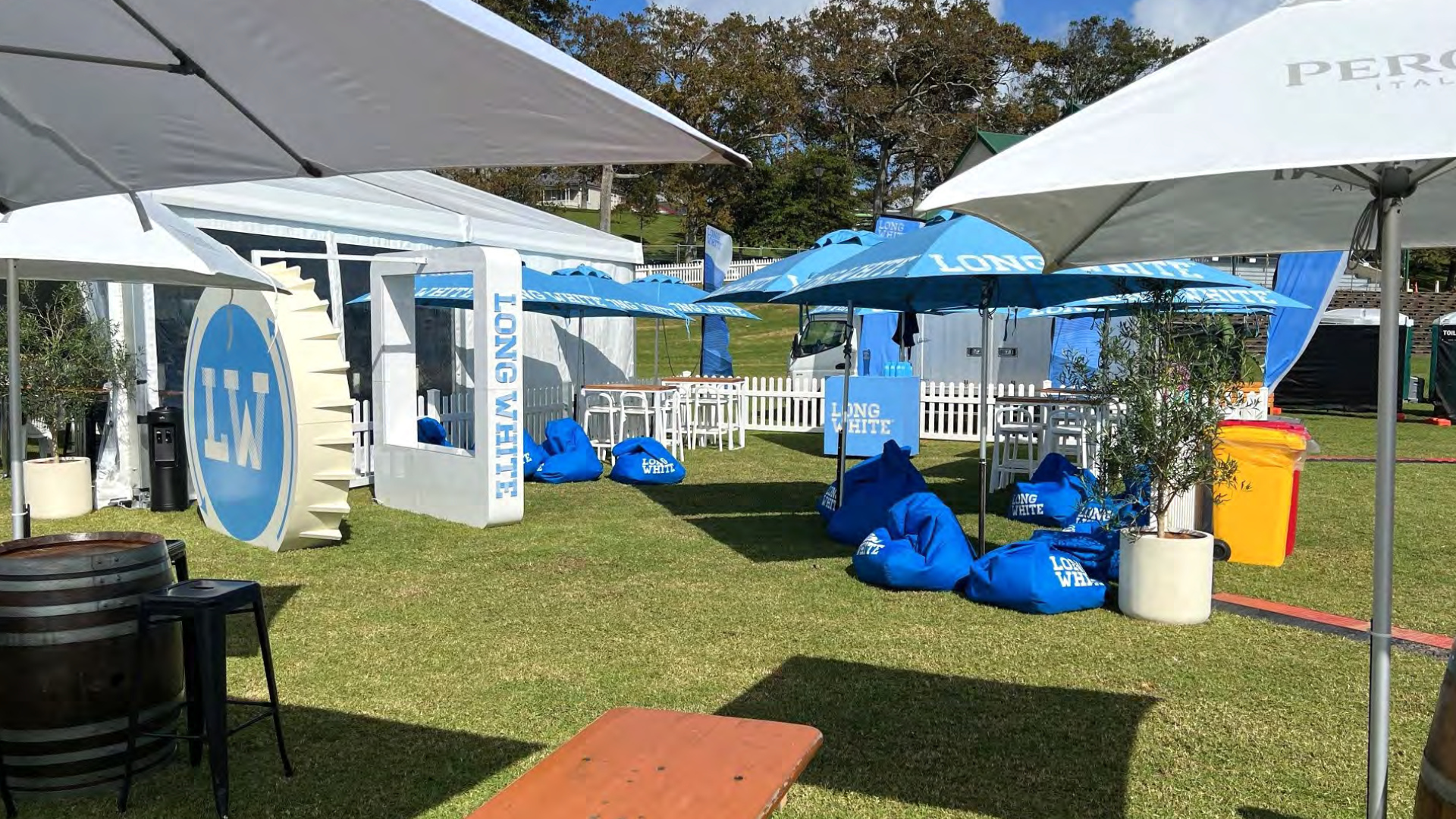 An outdoor event setup with white and blue umbrellas, tables, and chairs. There are blue Bean bags and signs with the text "Long White." The area is grassy with a white picket fence and trees in the background.