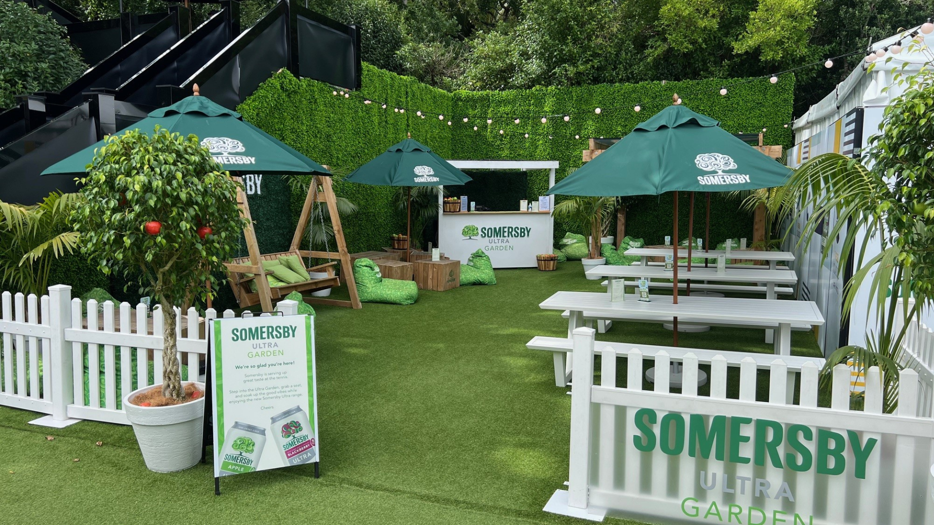 Outdoor garden setting with green umbrellas labeled 'SOMERSBY,' white picnic tables with umbrellas, a swing with green cushions, potted trees, and a small bar area in the background. Signage reads 'SOMERSBY ULTRA GARDEN' and promotional posters for Somersby drinks.