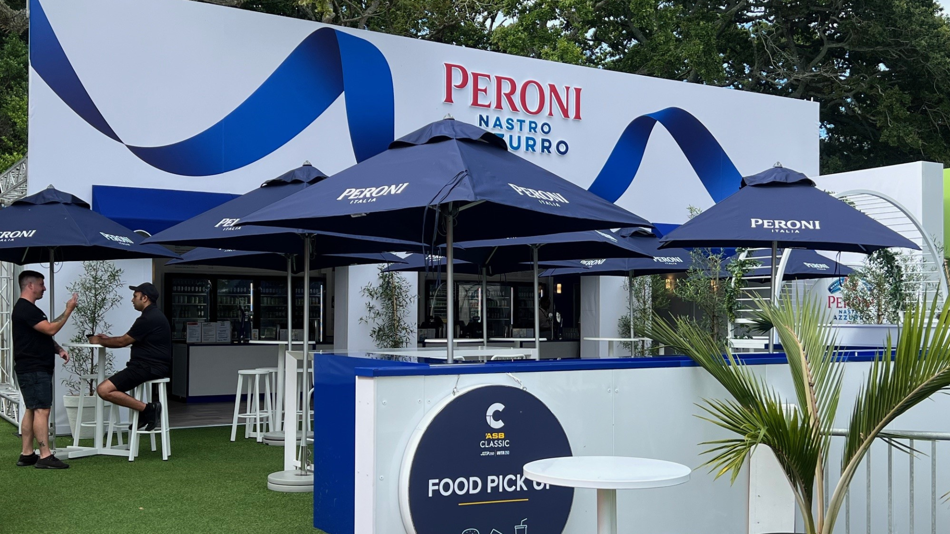 Outdoor food stand at a Peroni event with blue umbrellas, white seating, and a white and blue backdrop. Two people are talking, one sitting on a stool, with bar menus visible inside.